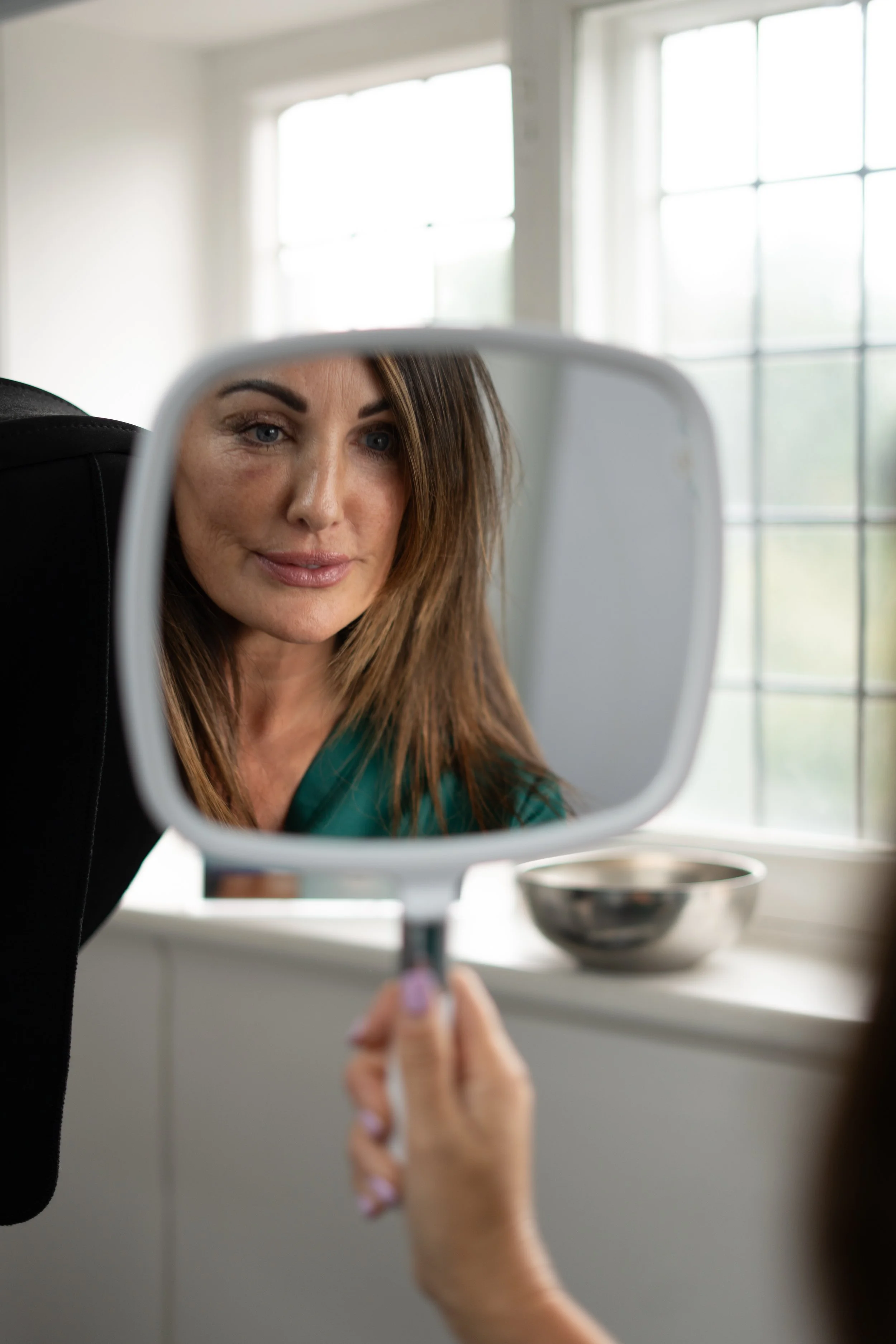 A woman looking into a handheld mirror, with her reflection visible, in a room with large windows.