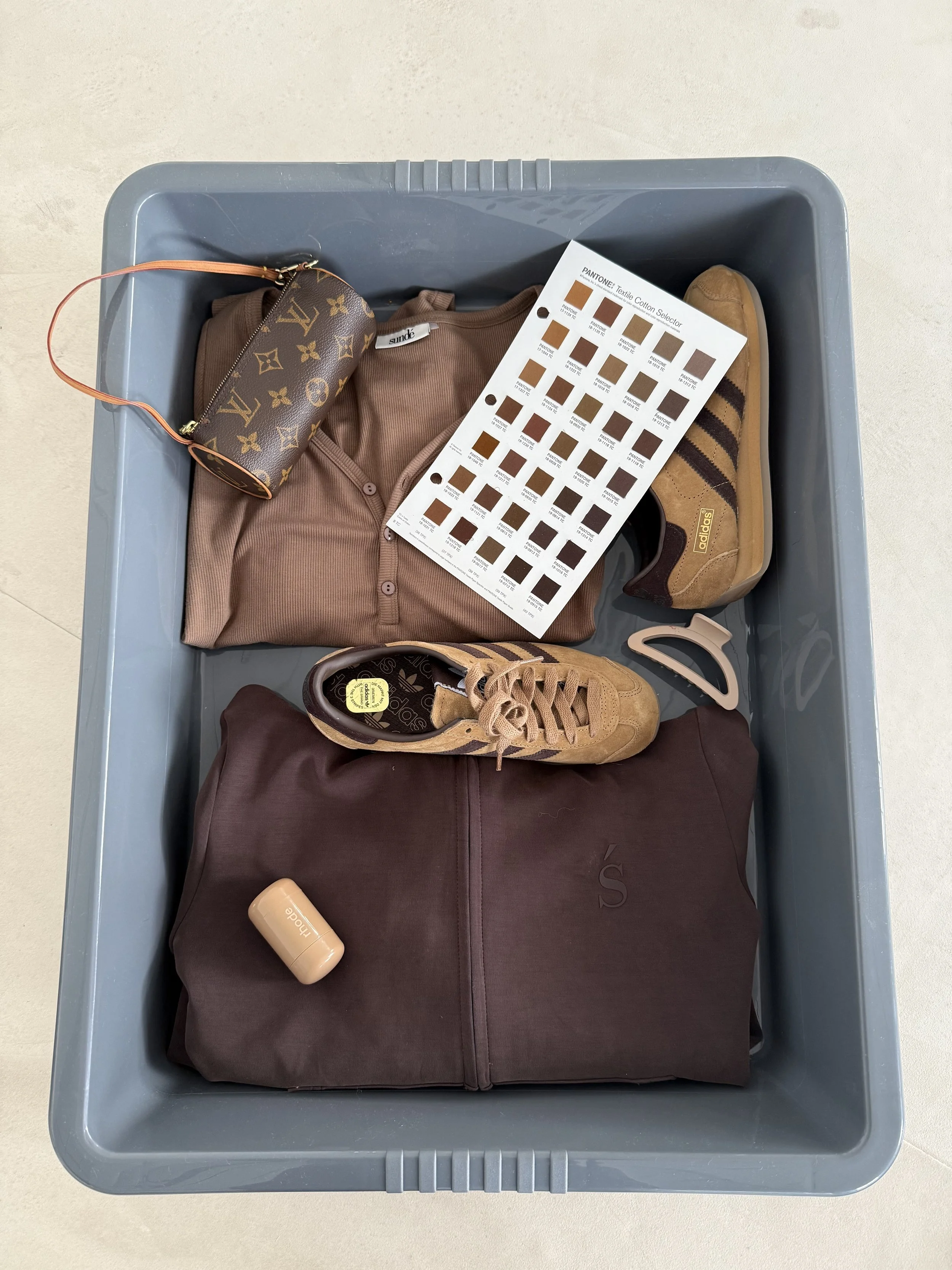 A gray plastic storage bin containing a folded brown shirt, a yellow sneaker, a maroon jacket, a small Louis Vuitton clutch, a beige AirPod case, a color swatch, and a brown sneaker.