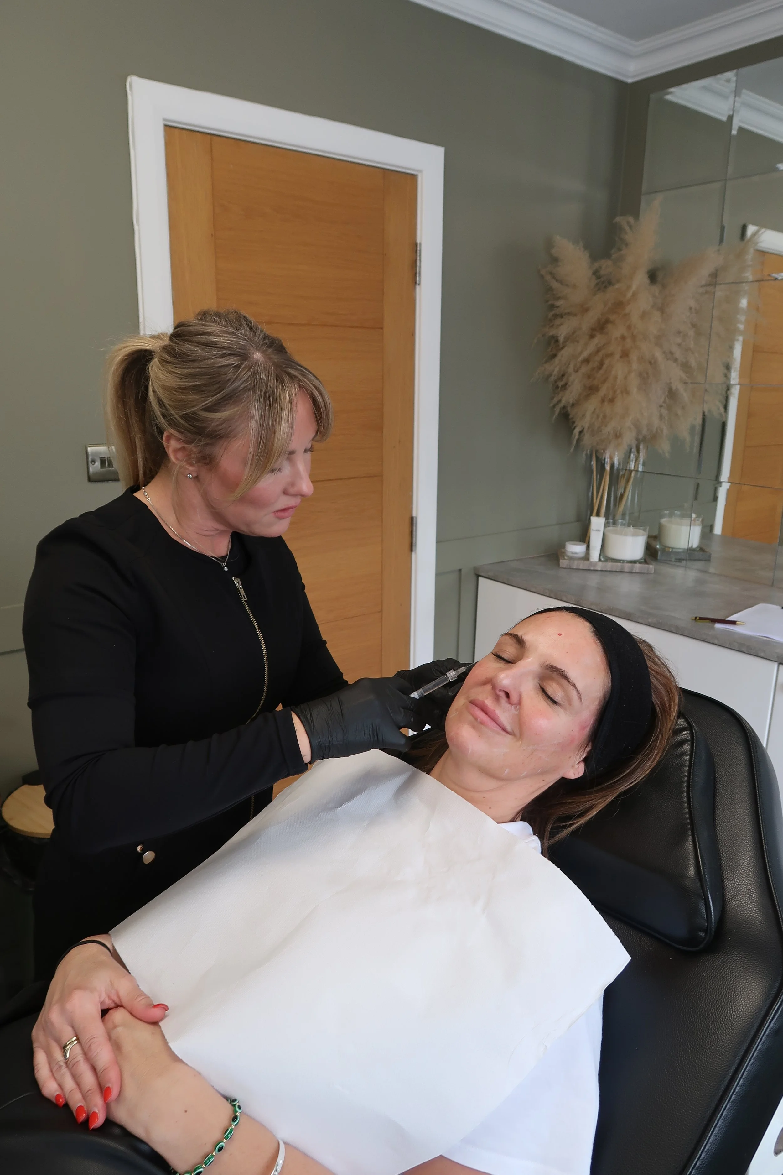 A woman with light brown hair and a black headband is lying in a chair with her eyes closed, receiving a cosmetic treatment. A second woman with blonde hair tied back and wearing black gloves is performing the procedure.