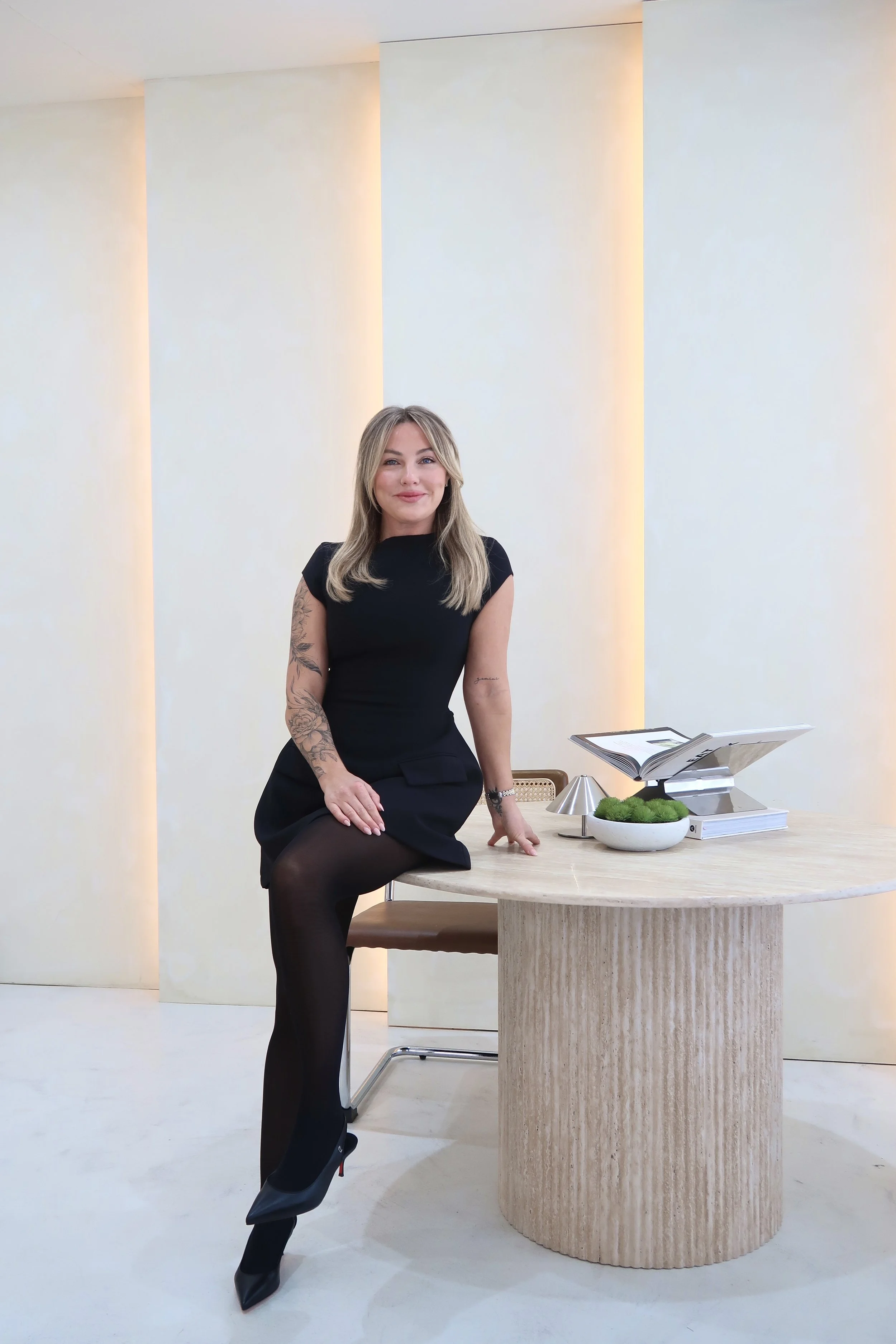 A woman with blonde hair and tattoos on her arm, dressed in a black dress and black tights, sitting at a round wooden table in a minimalistic, well-lit room with white walls. The table has a bowl of green moss and a stack of papers or books, with a music stand nearby.