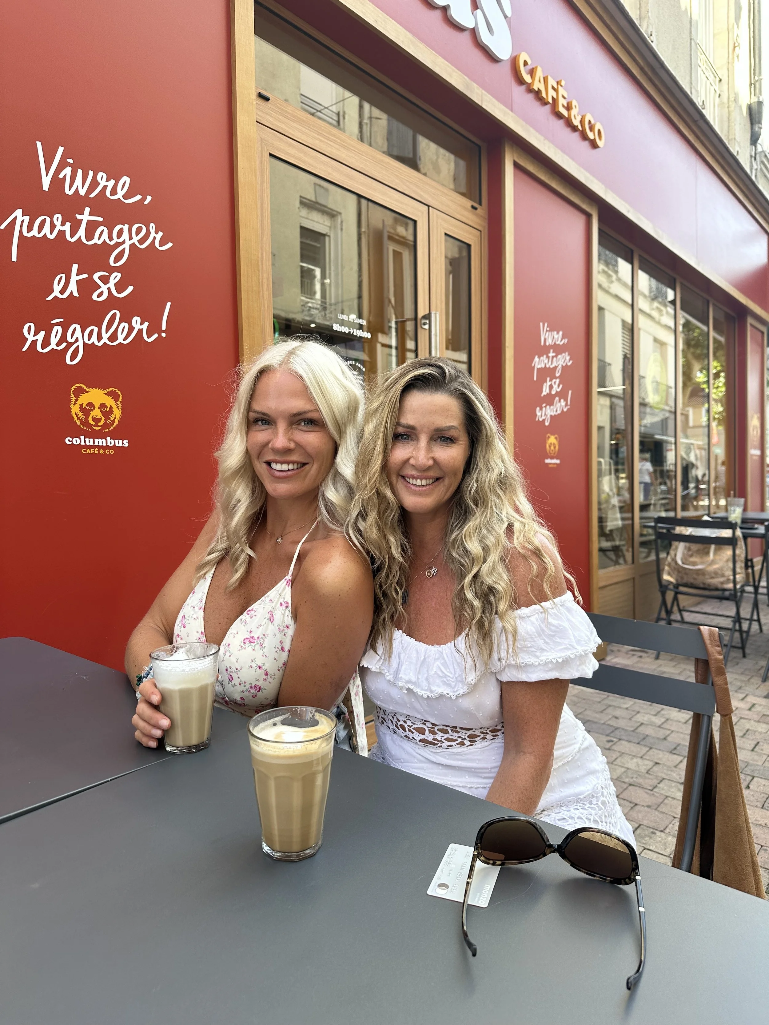 Two women sitting at an outdoor café table, smiling, with drinks in front of them. A pair of sunglasses is on the table. The café has a red exterior wall with French writing and a logo featuring a lion's face.