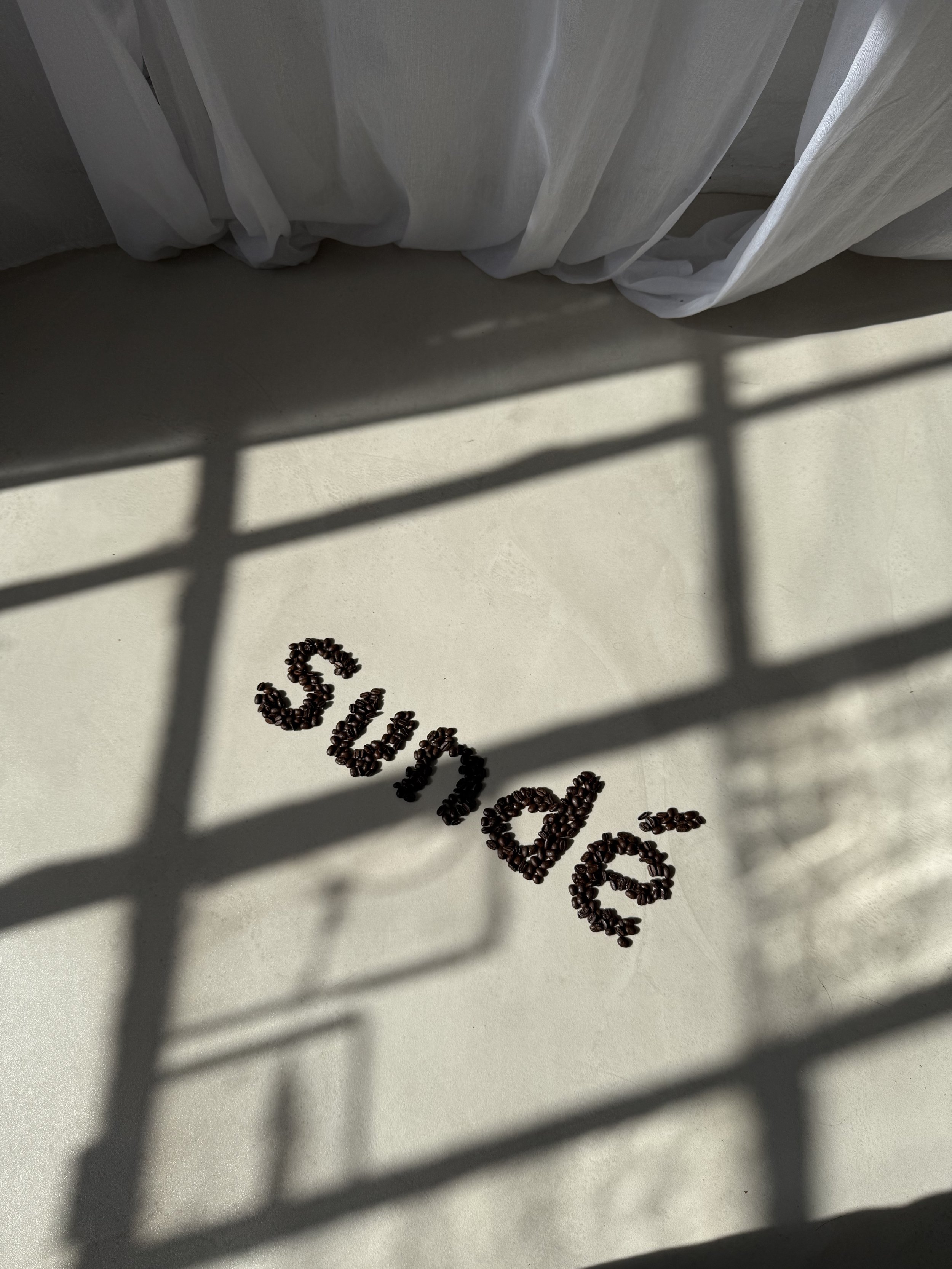 The word 'sleep' spelled out with coffee beans on a light-colored floor, with shadows cast by a window and a white curtain in the background.