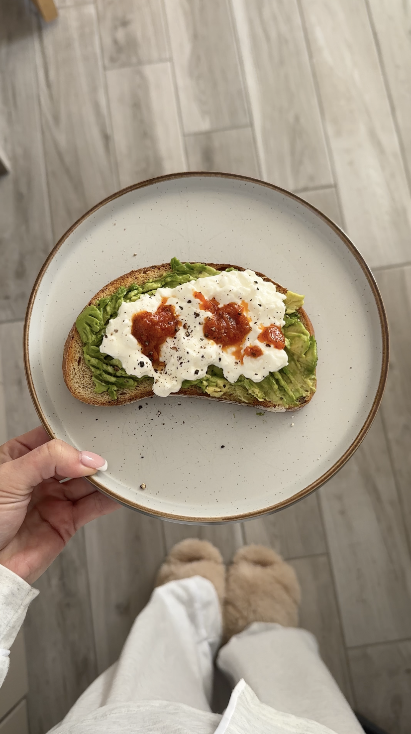 A plate with a slice of toast topped with lettuce, white sauce, red sauce, and black pepper, held by a person's hand, with a gray tile floor and fuzzy slippers in the background.