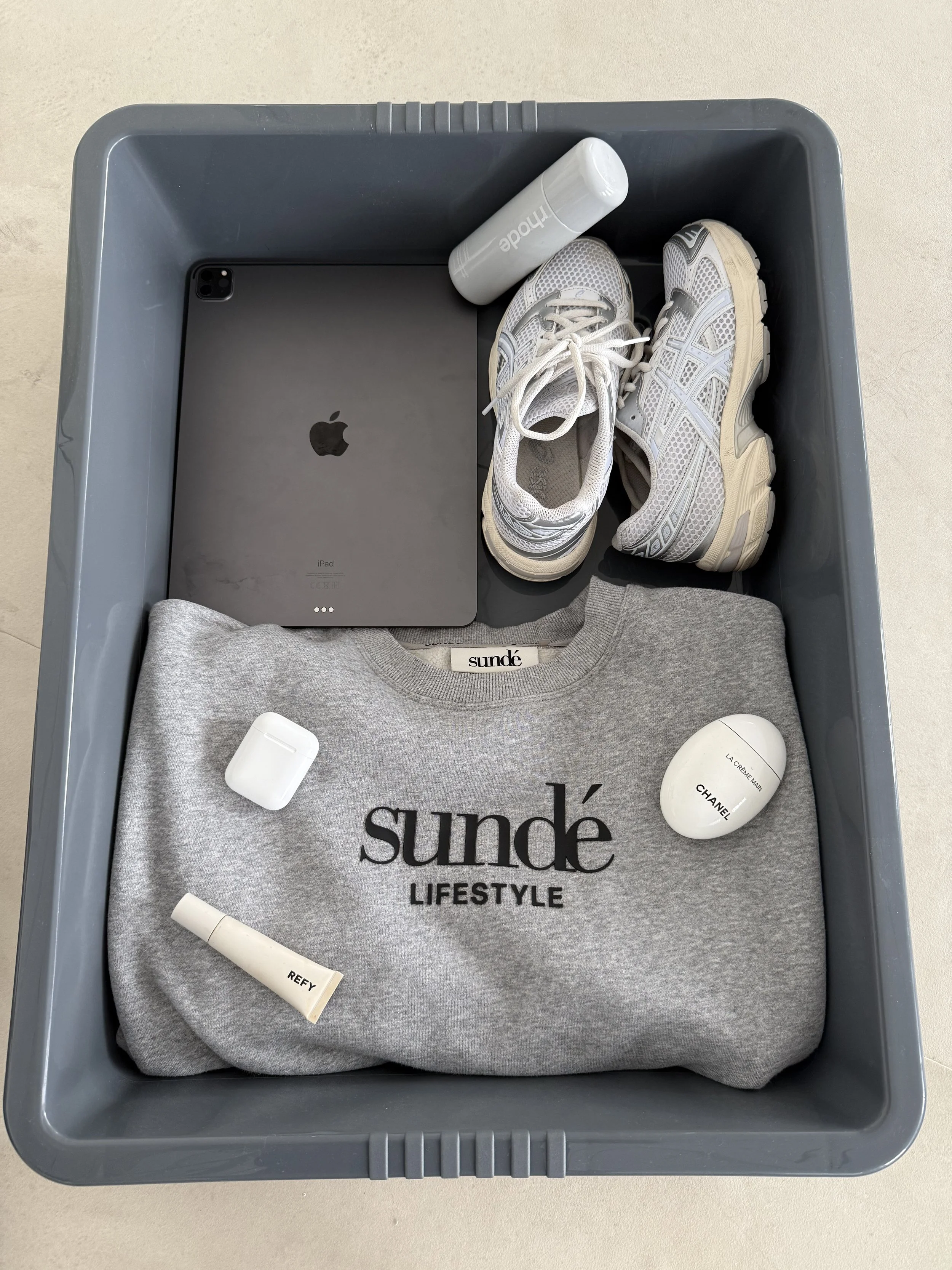 A gray plastic bin containing a folded gray sweatshirt with 'sundé LIFESTYLE' printed on it, a pair of white sneakers, a gray iPad, a small white bottle of perfume, a white AirPods case, a white cylindrical container labeled 'Refy', and a white Chanel perfume bottle, all arranged on top of a light-colored surface.
