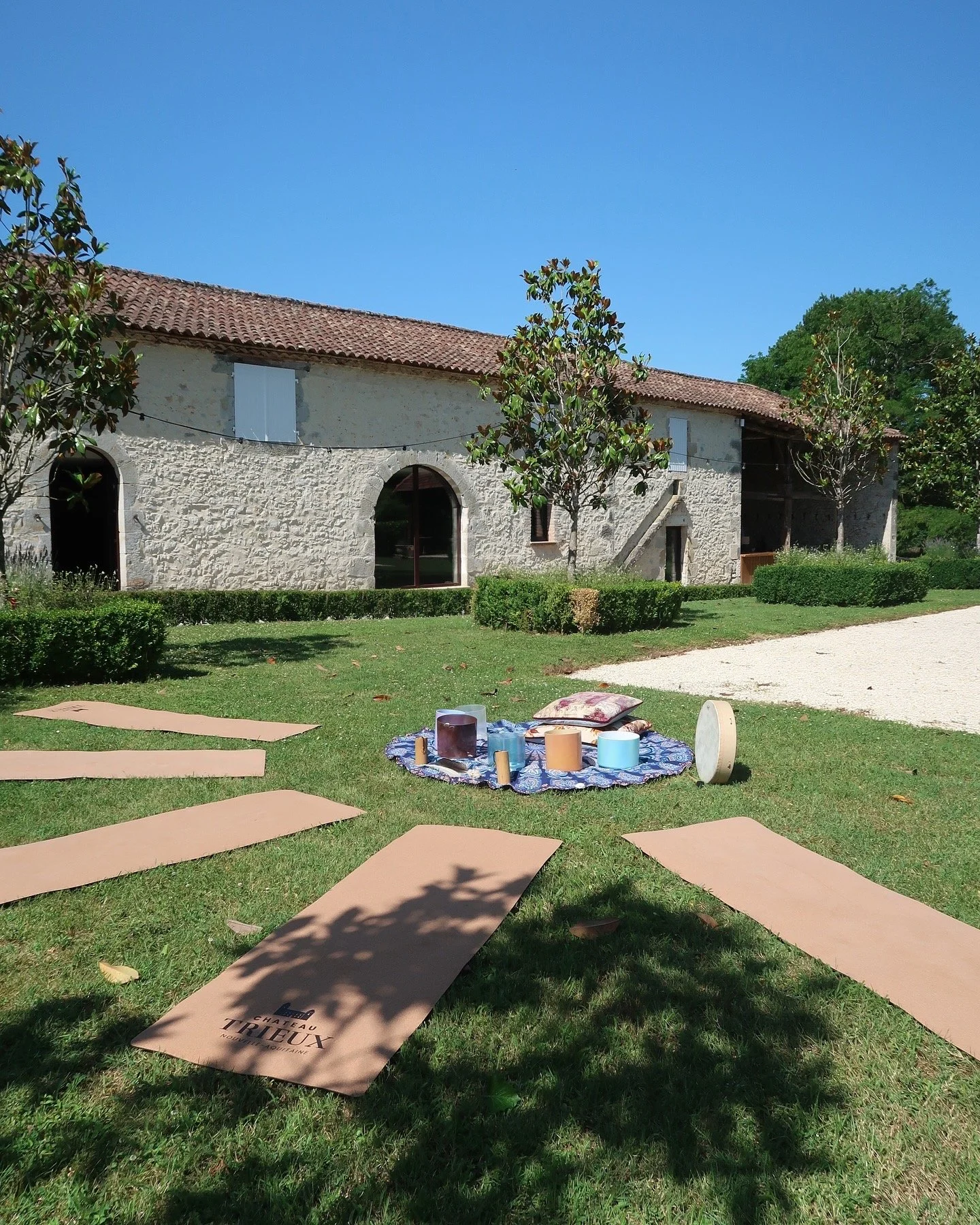 Yoga mats arranged outdoors on grass with a circular setup of candles and pillows, with a stone house and trees in the background on a sunny day.
