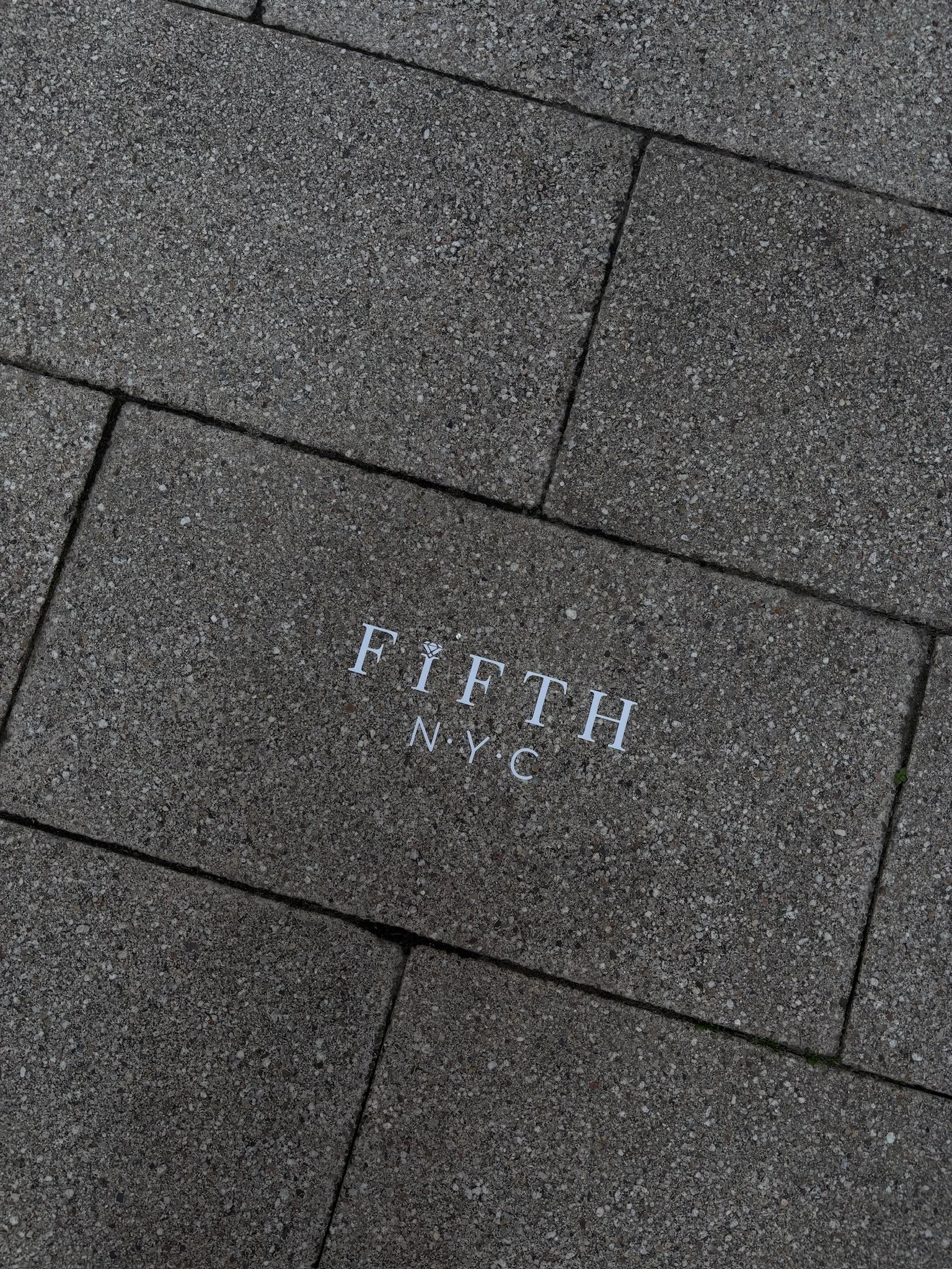 A stone sidewalk with a tile engraved that reads "FIFTH N.Y.C." in white letters.