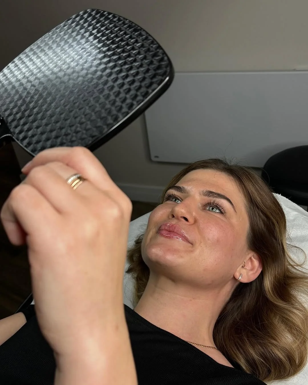 A woman lying on a medical bed with a cardboard facial mask on her face, holding a handheld mirror and looking at her reflection.