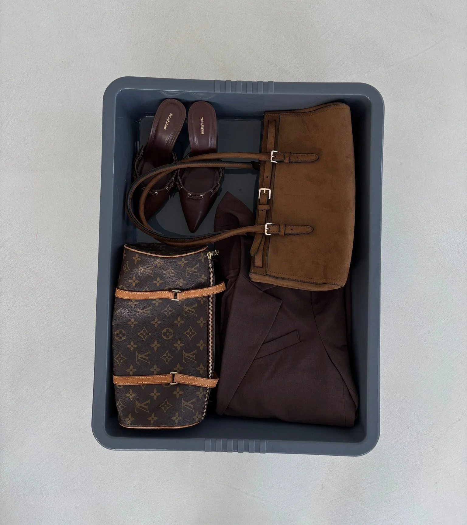 A plastic storage bin containing brown high-heeled shoes, a brown suede handbag, a Louis Vuitton patterned duffel bag, a dark brown blazer, and a brown belt.