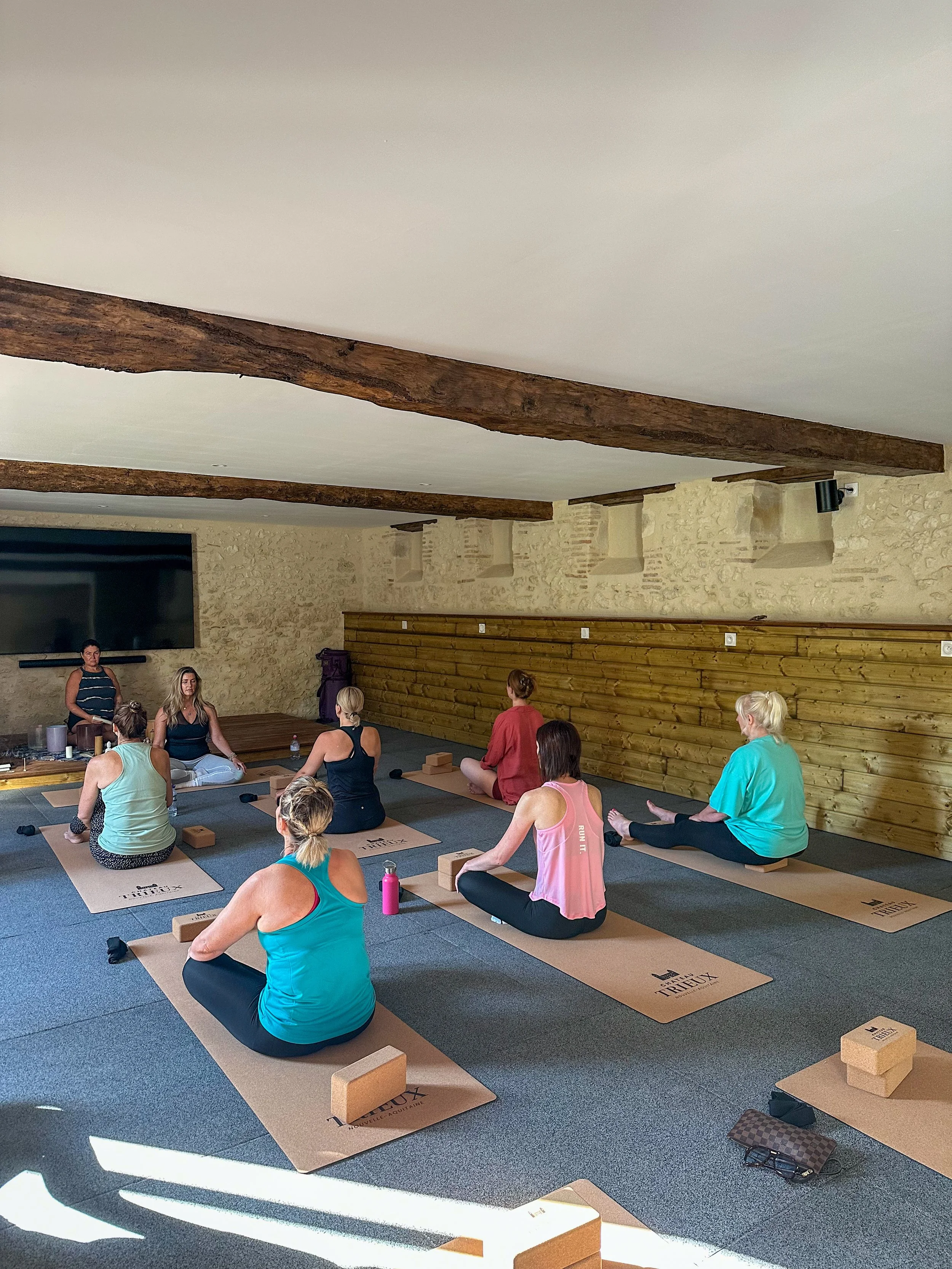 A group of women participating in a yoga class in a rustic studio with exposed wooden beams and stone walls, seated on mats with yoga blocks and water bottles.