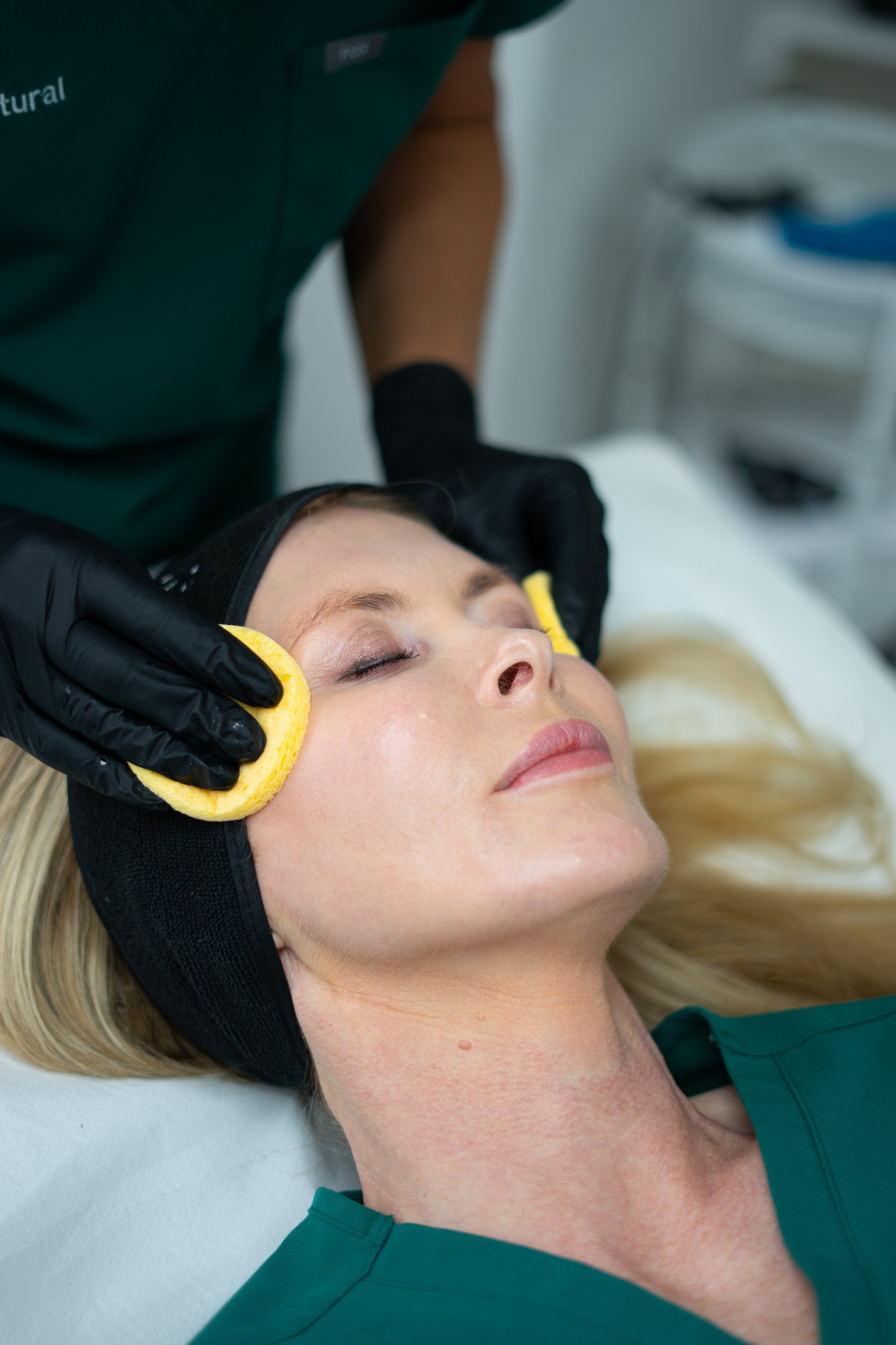 A woman lying down with eyes closed, receiving a facial treatment with a yellow sponge on her face, attended by a practitioner wearing black gloves in a clinical setting.