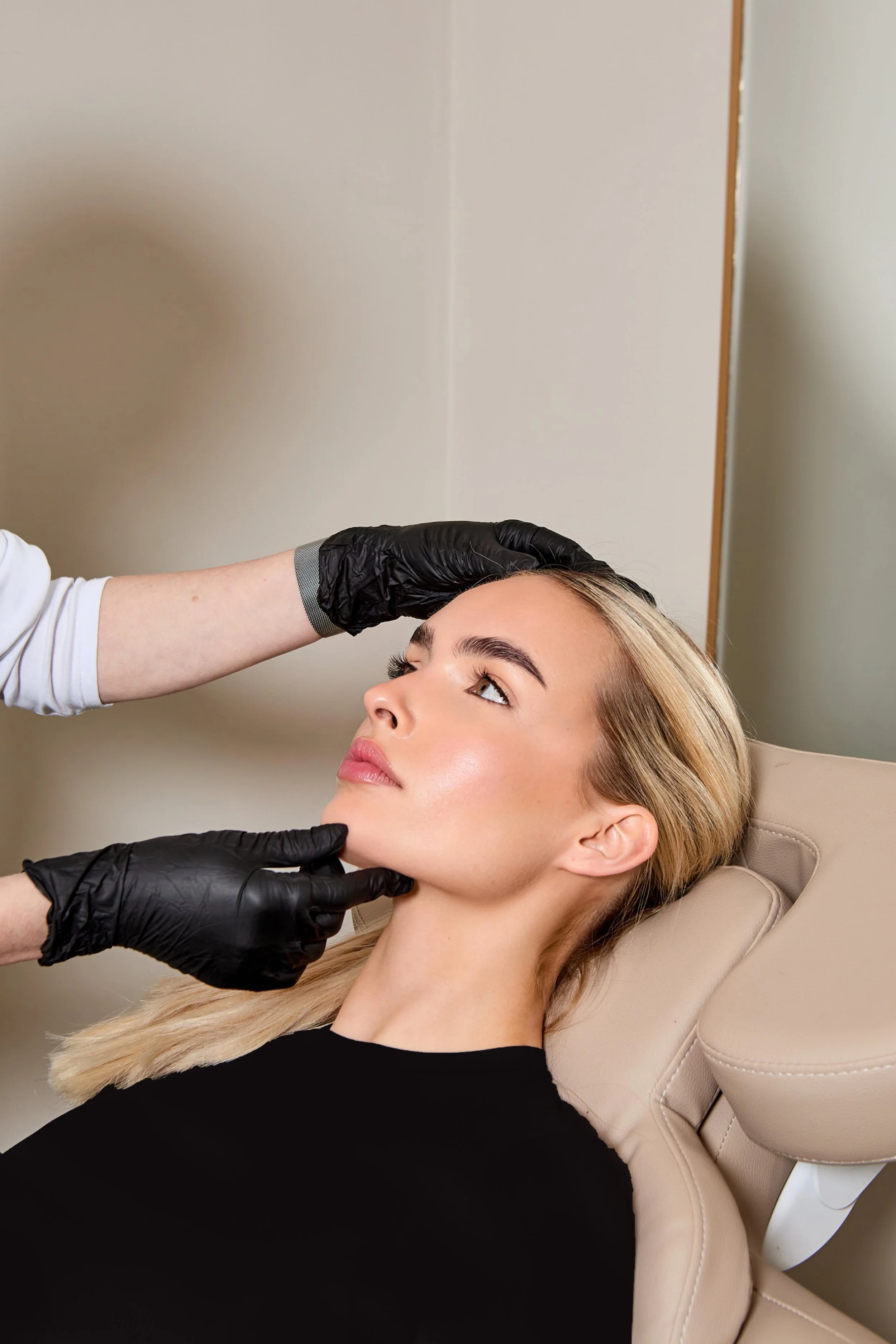 A woman is lying back in a chair at a medical or cosmetic clinic, with her head resting on a headrest. A healthcare professional wearing black gloves is examining her forehead and chin.