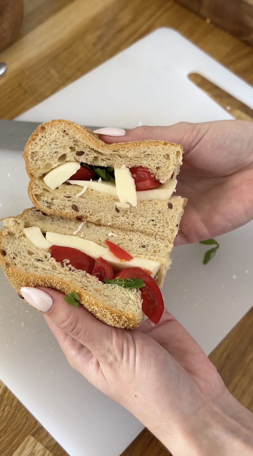 Close-up of hands holding a sandwich with tomato, cheese, basil, and seeds on multigrain bread.