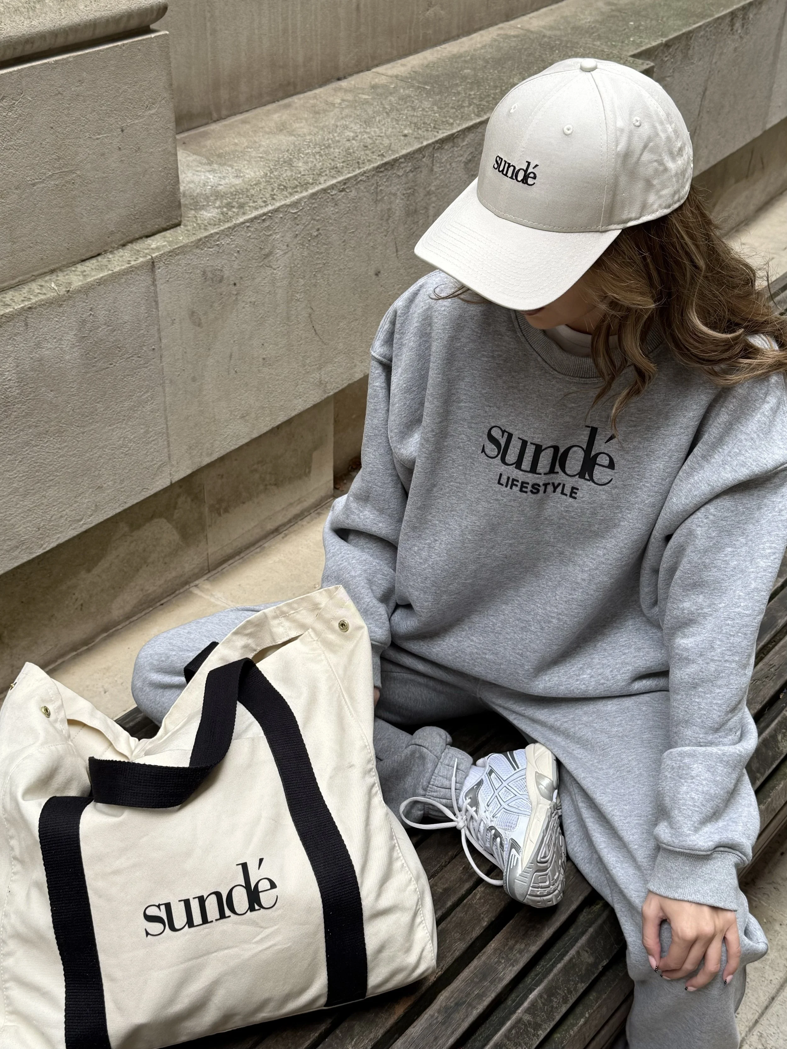 A woman sitting on a wooden bench wearing a gray sweatshirt, gray sweatpants, a white cap, and white sneakers. She has a beige bag with black straps next to her, both with the word "sunde" printed on them.