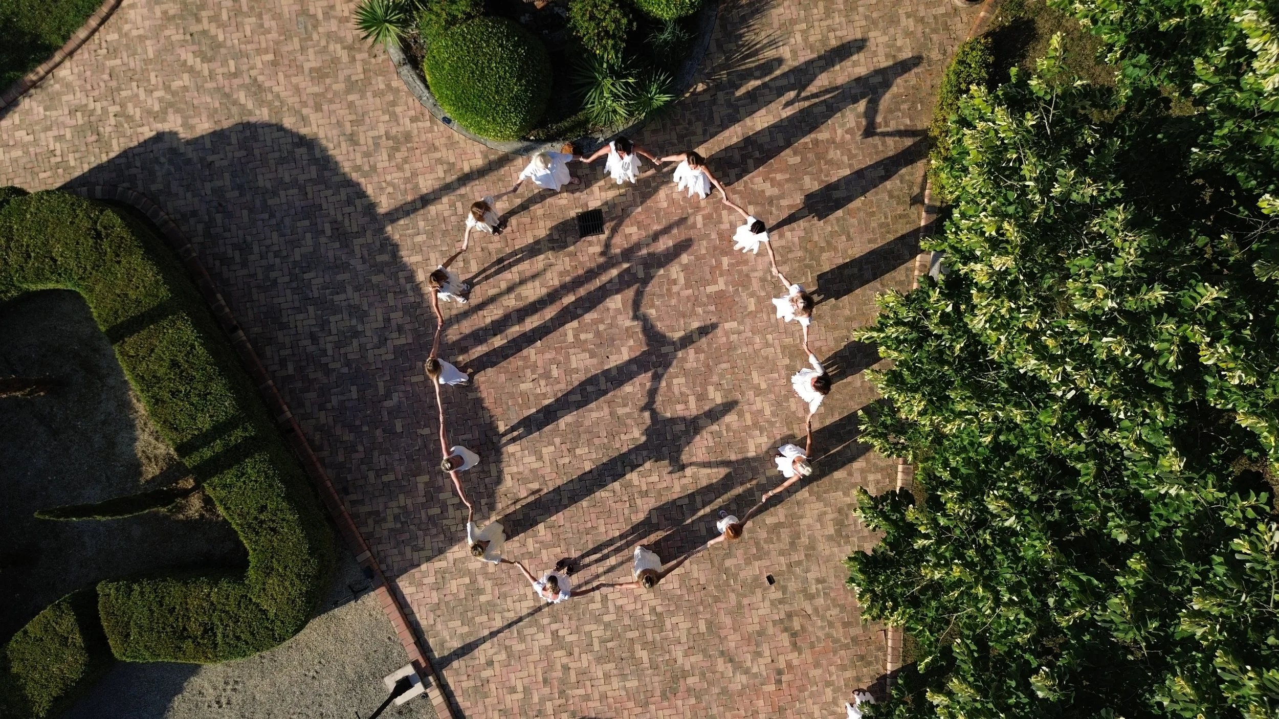 A group of children dressed in white holding hands forms a heart shape on a brick pathway outdoors, with shadows cast by trees and the children.