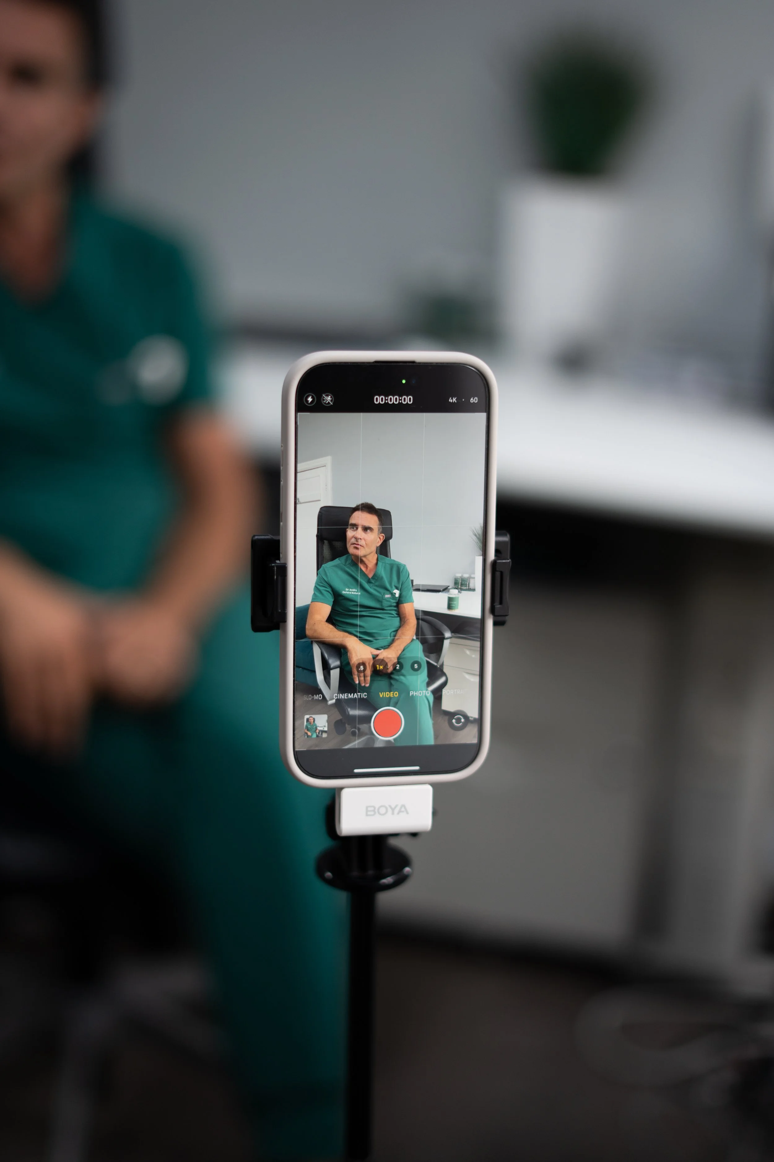 A smartphone on a stand recording a man in medical scrubs sitting in a chair in an office.