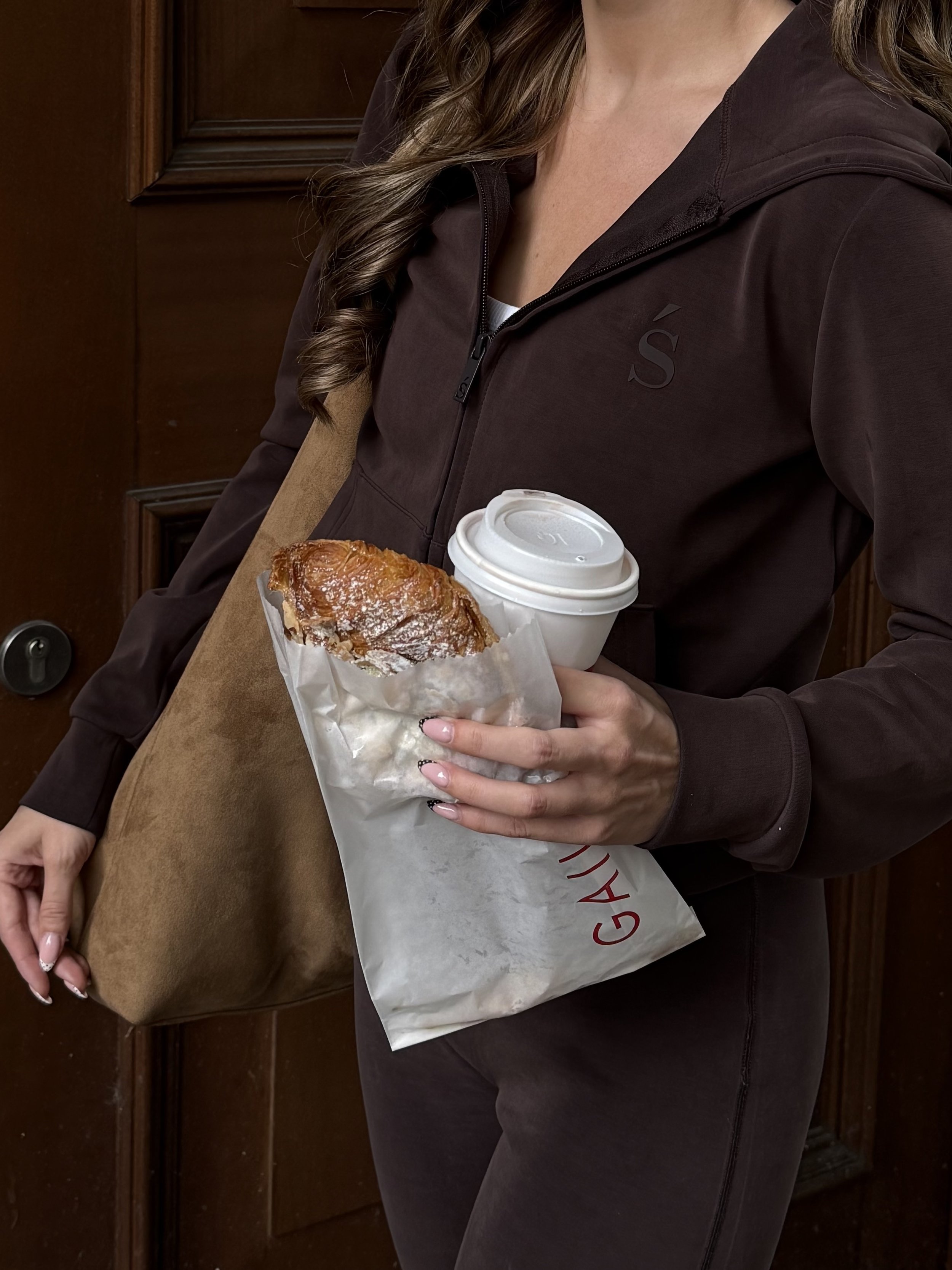 A woman wearing a brown jacket holding a sandwich and a disposable coffee cup, with a brown tote bag hanging from her shoulder.