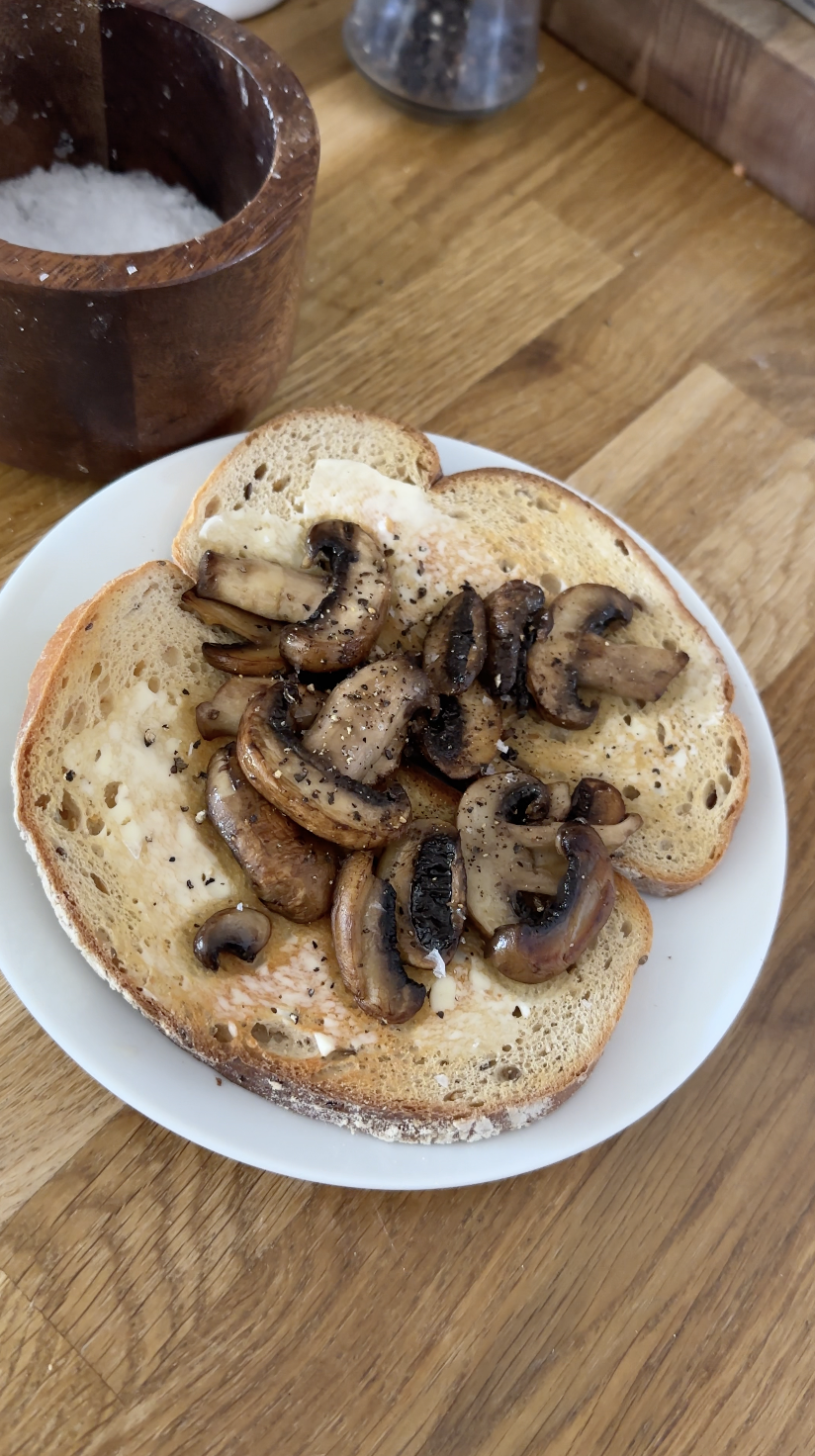 Slices of toasted bread topped with sautéed mushrooms and melted butter, on a white plate. In the background, a wooden container of salt and a glass container, all on a wooden table.