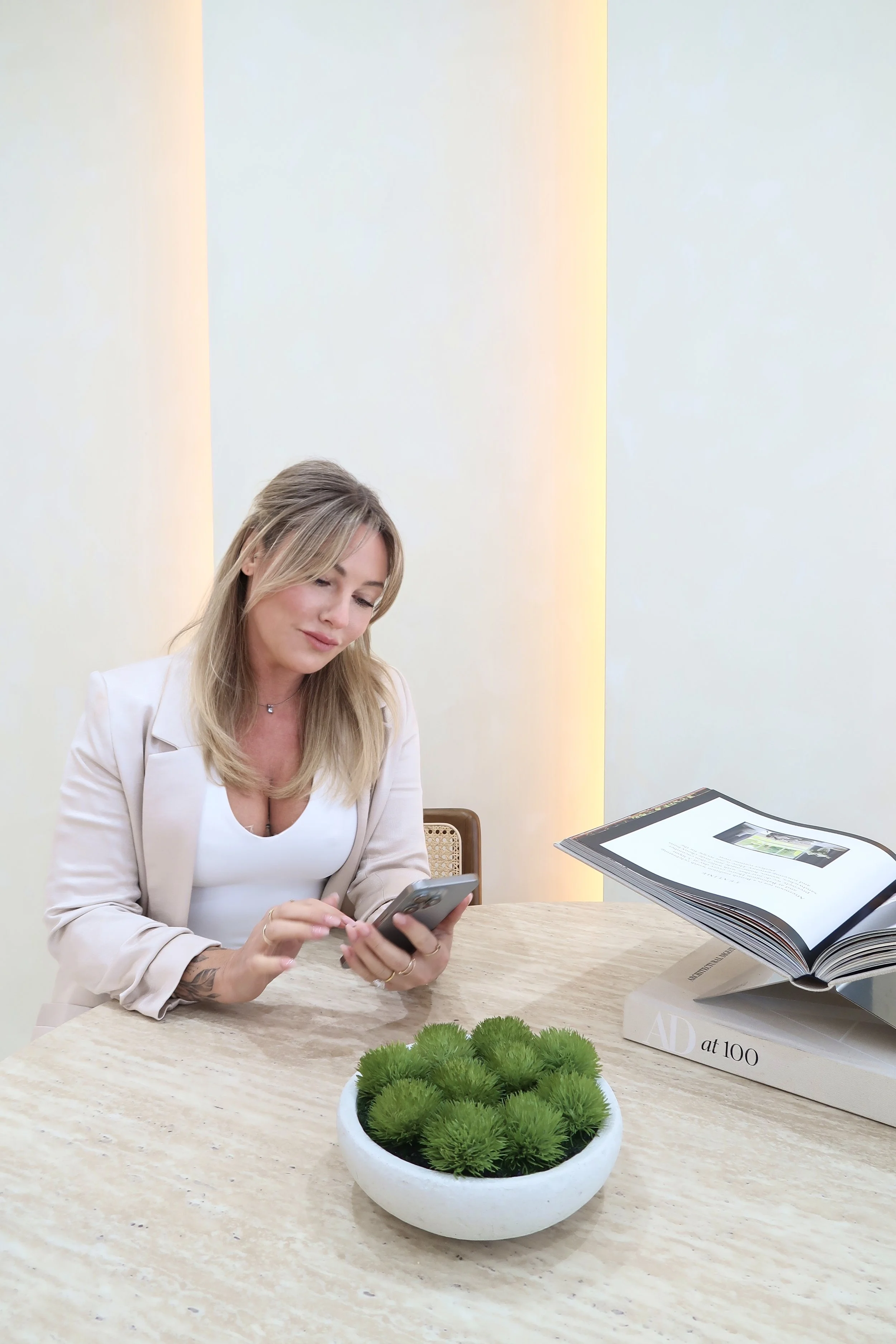 A woman with blonde hair, wearing a beige blazer and white top, sitting at a table in an office or meeting room, looking at her phone. There is a plant in a white pot, a stack of large books, and an open magazine on the table, with soft lighting on the wall behind her.