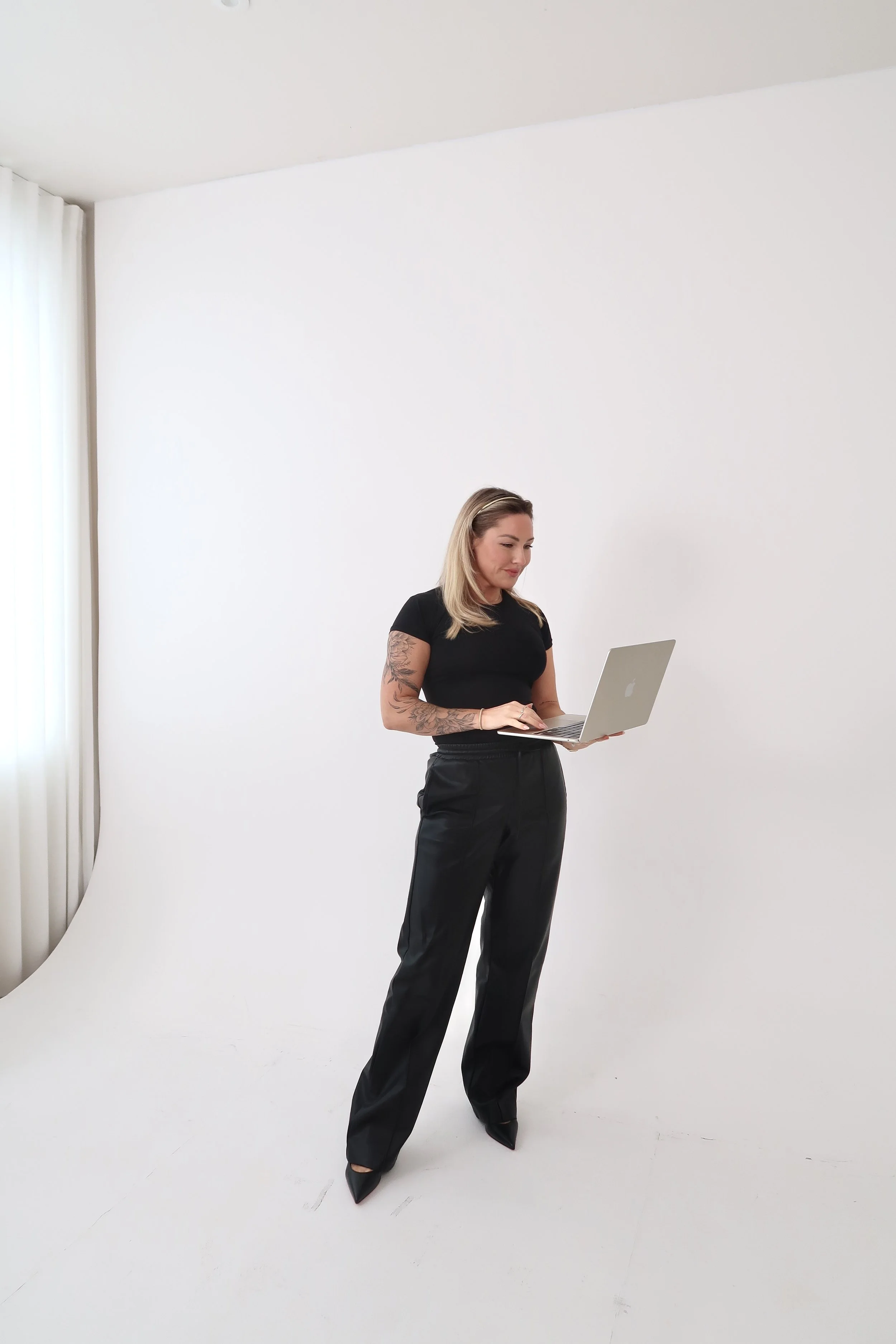 A woman with blonde hair, tattoos on her right arm, wearing a black T-shirt, black pants, and black pointed shoes, standing in front of a white seamless background while looking at a silver laptop.