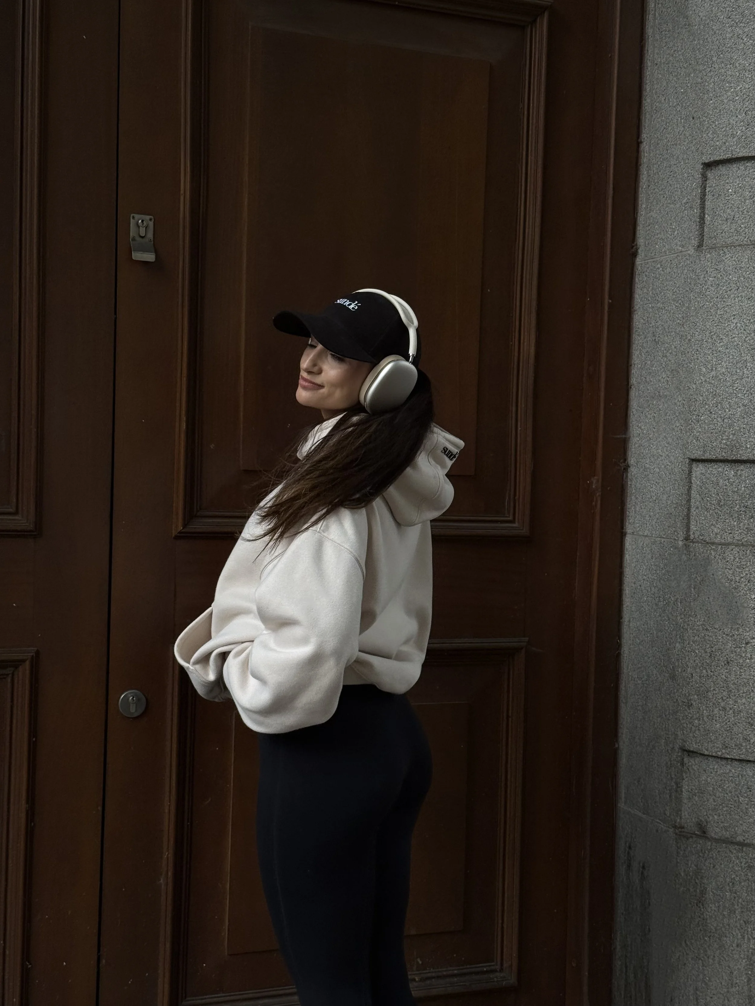 A woman wearing a black cap, beige hoodie, and black leggings listening to music with beige headphones, standing in front of a wooden door and a gray stone wall.