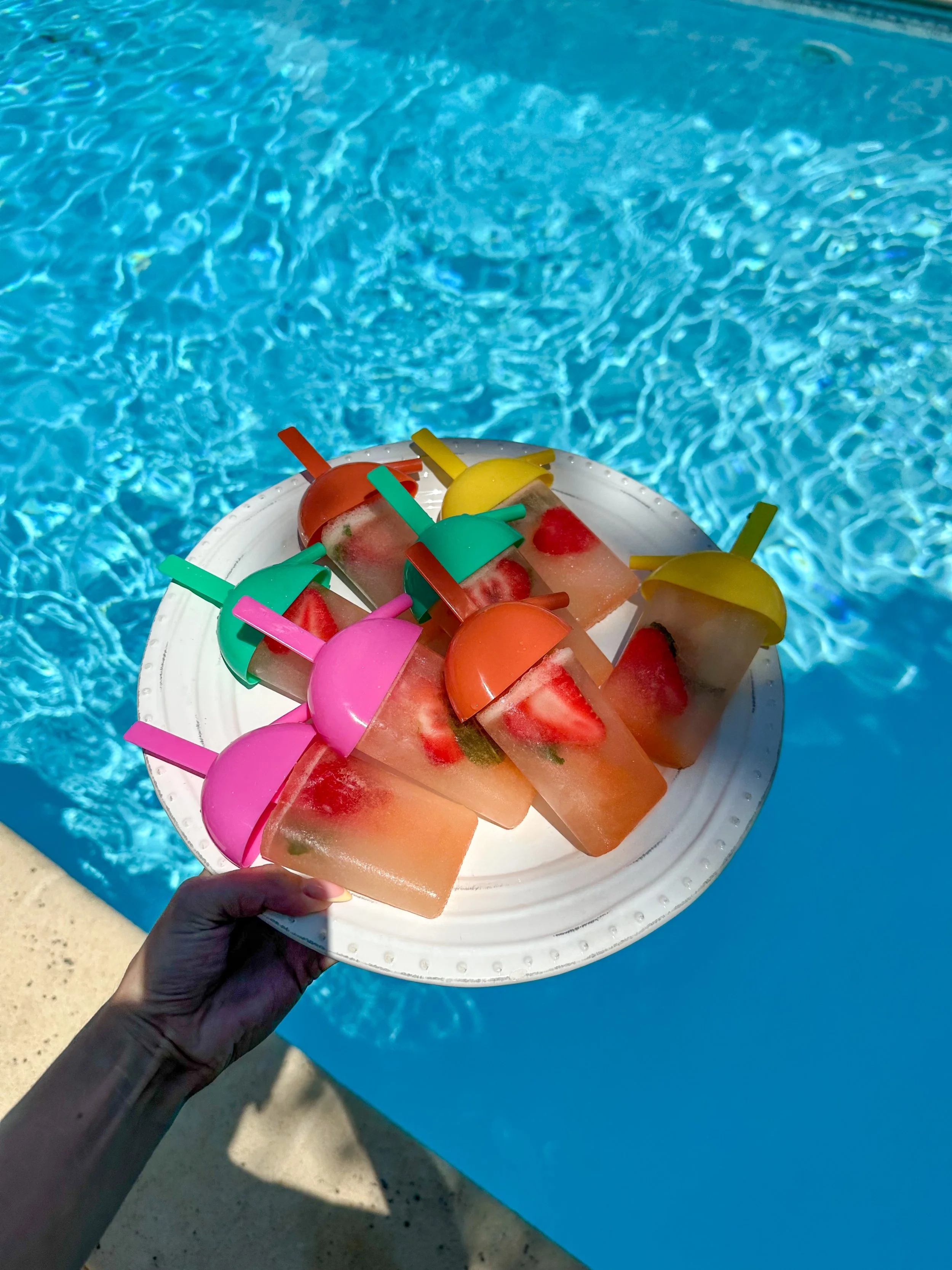 A person's hand holding a white plate with popsicles shaped like watermelons, featuring slices of strawberry and lemon slices as toppings, against the backdrop of a swimming pool with clear blue water.
