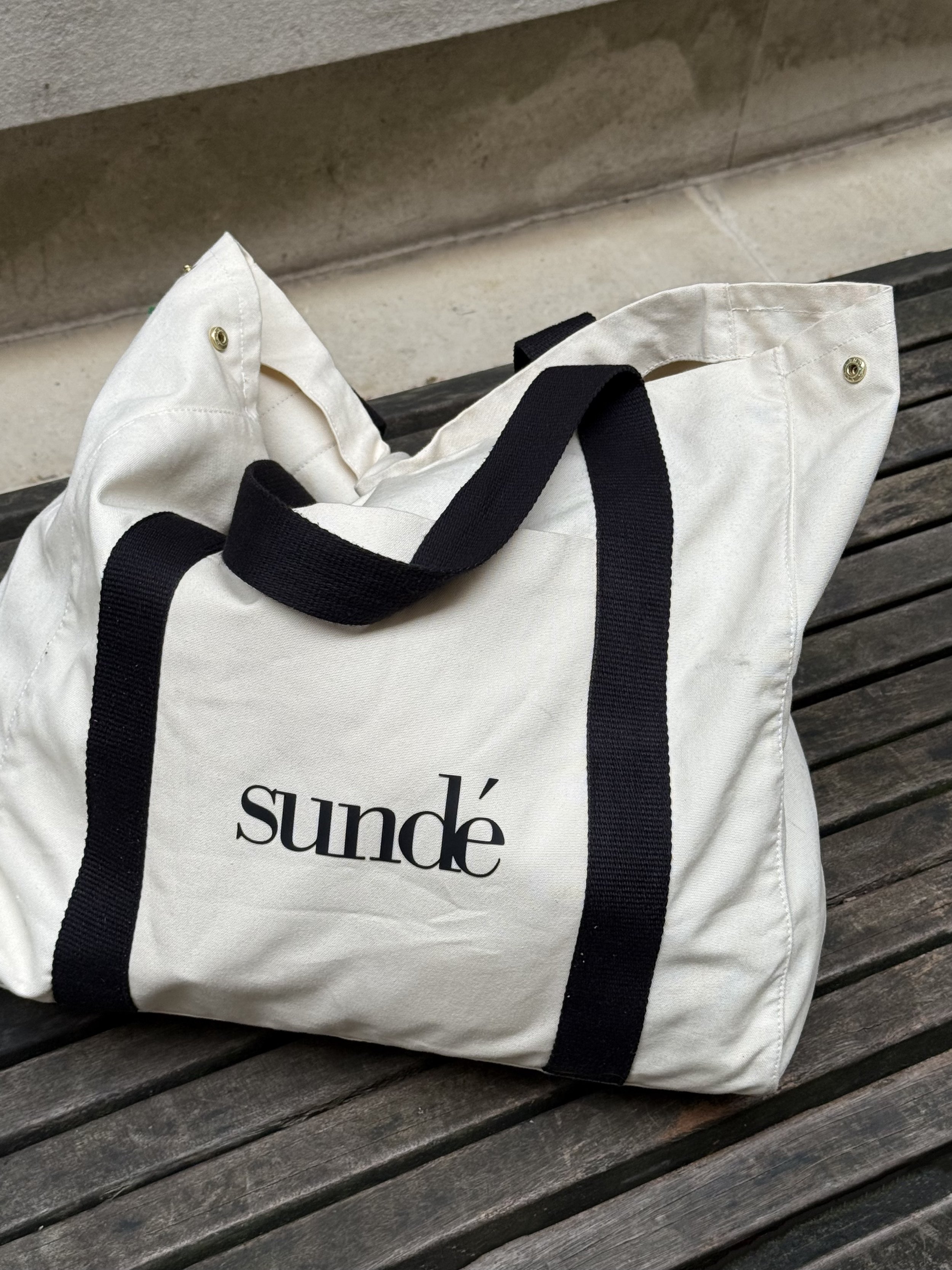 A white tote bag with black straps and the word 'sundé' printed on it, resting on a wooden bench.