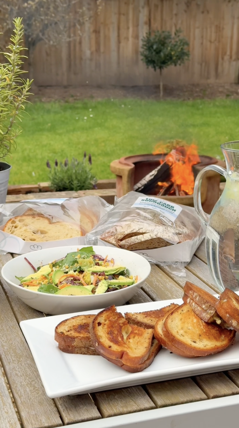 Outdoor table with toasted bread, fresh salad, and packs of bread in a backyard with a small fire pit and green grass.