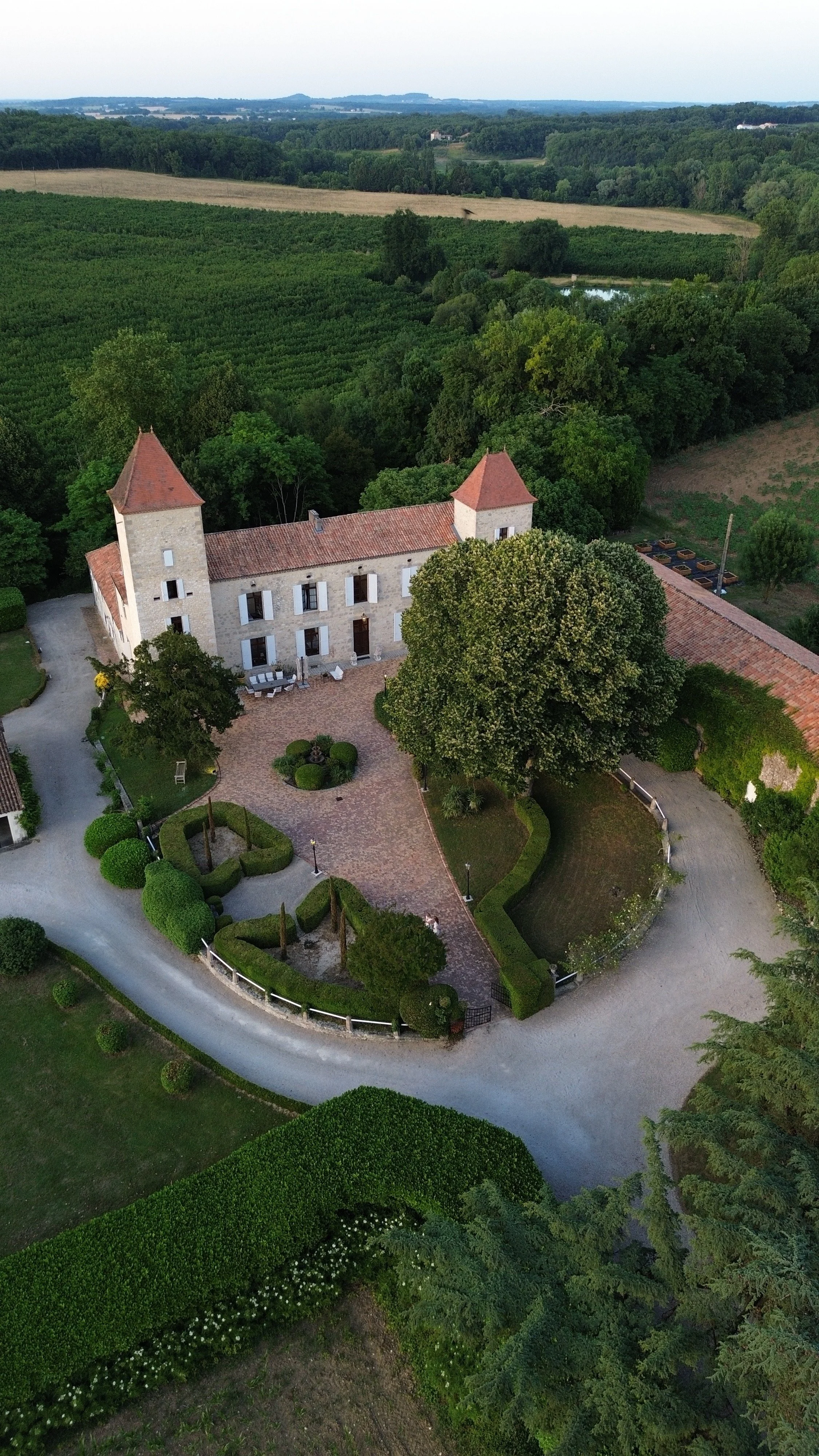 Aerial view of a historic stone castle with towers, surrounded by well-maintained gardens, trees, and farmland in the distance.
