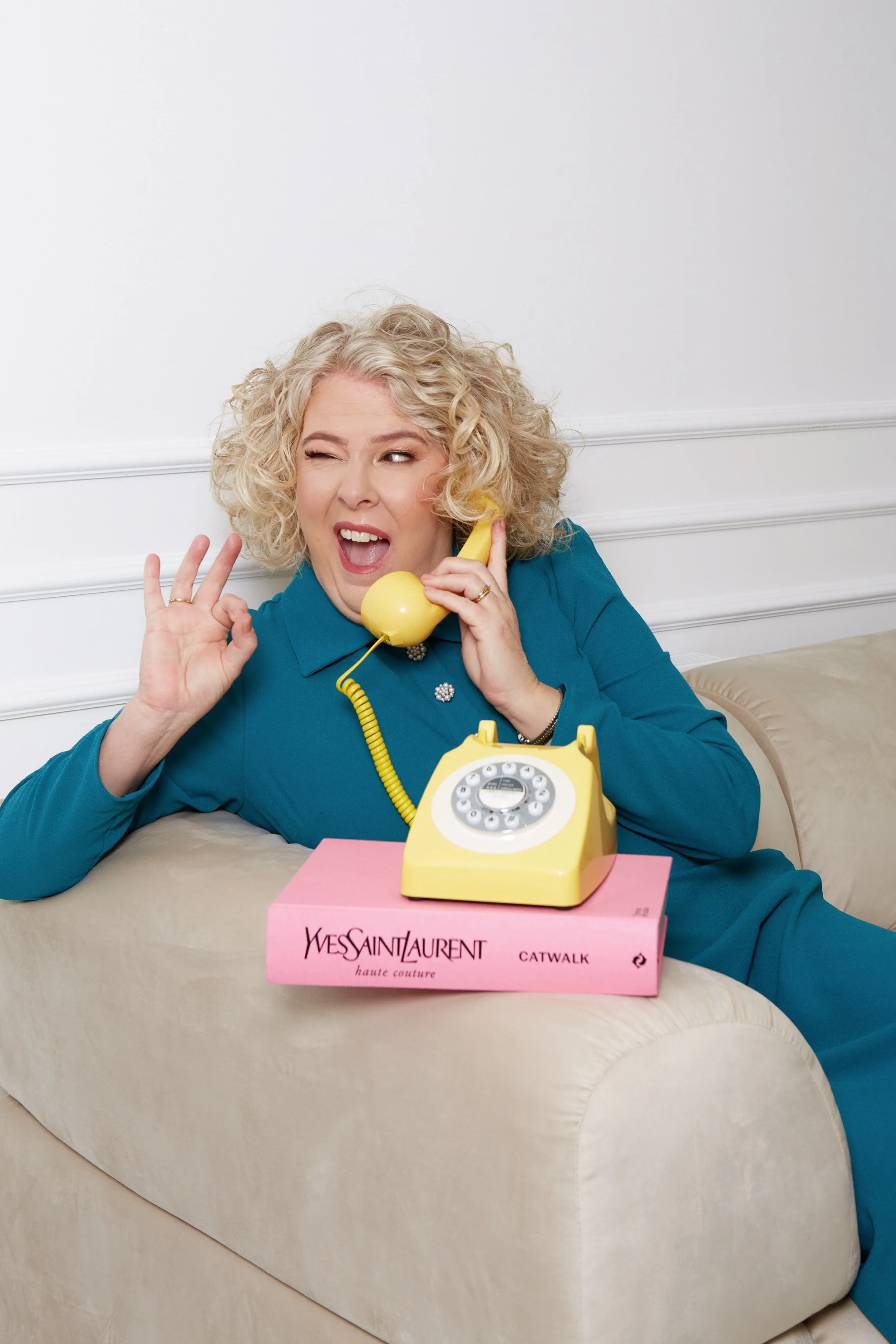 A woman with curly blonde hair sits on a beige couch, speaking on a yellow rotary phone, with a pink Yves Saint Laurent box labeled 'Catwalk' in front of her, making a playful facial expression.
