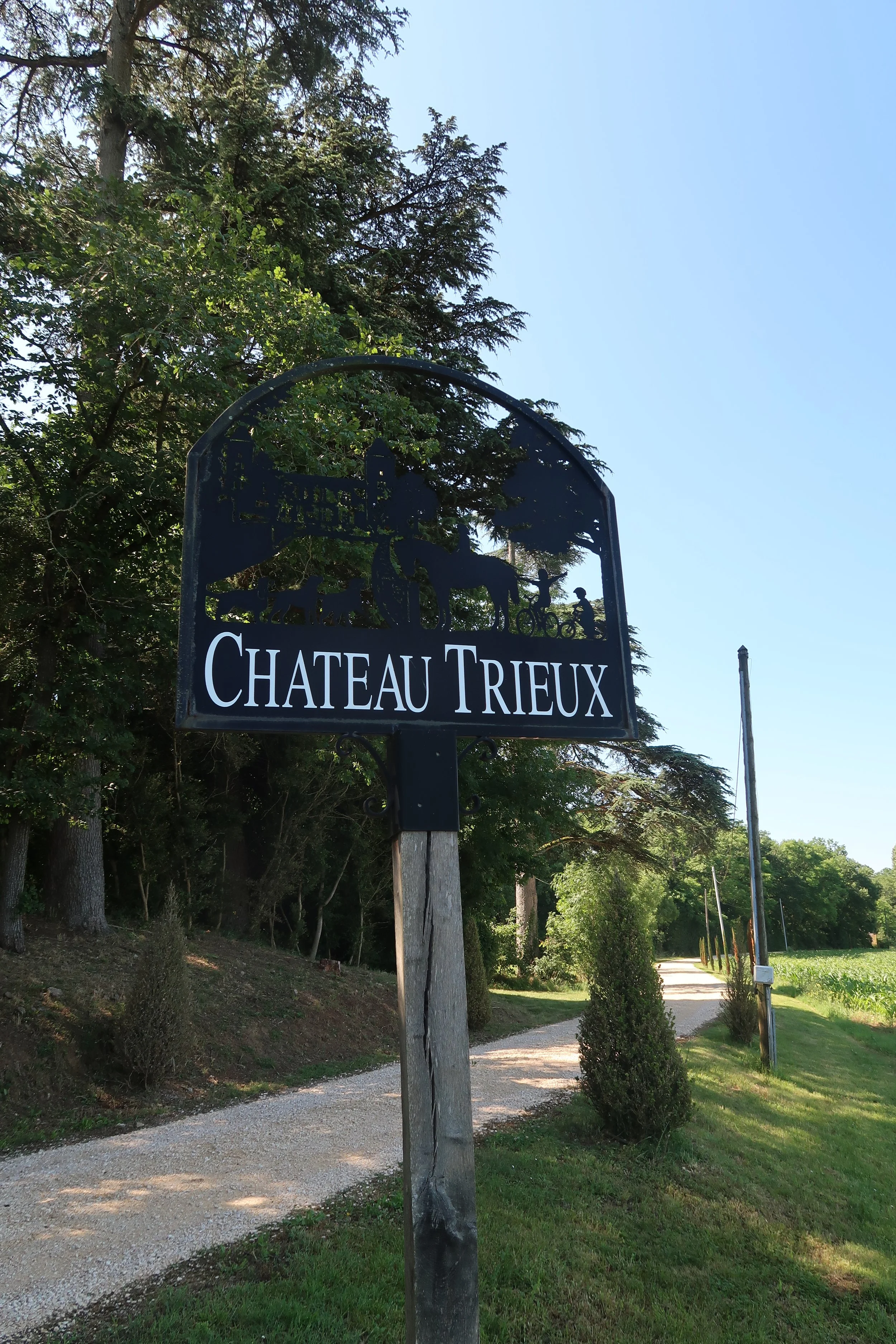 Entrance sign for Chateau Trieux featuring illustrations of a castle, trees, and people on bicycles, with a gravel pathway and greenery in the background.