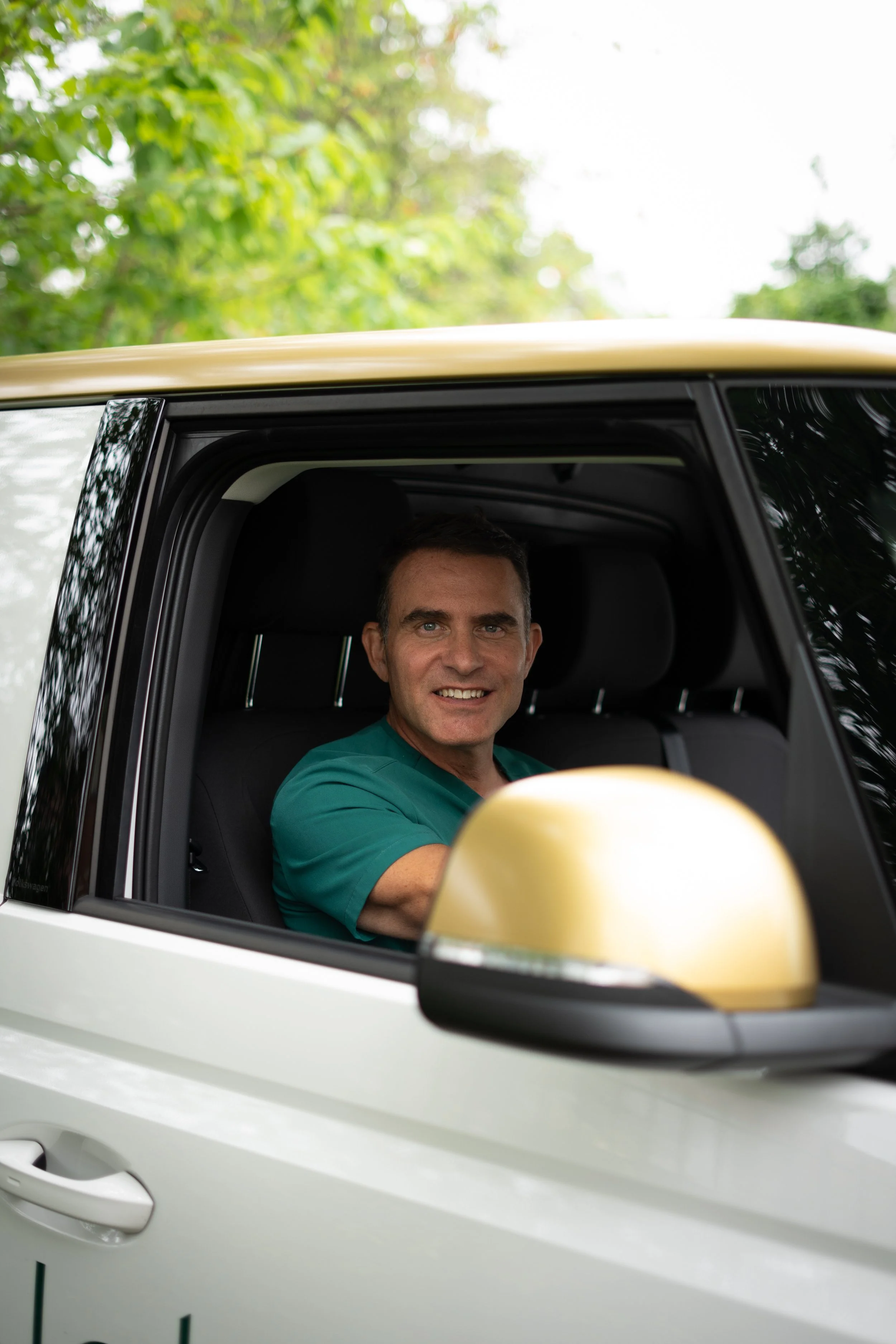A smiling man in medical scrubs sitting inside a white vehicle with the window rolled down, surrounded by trees.