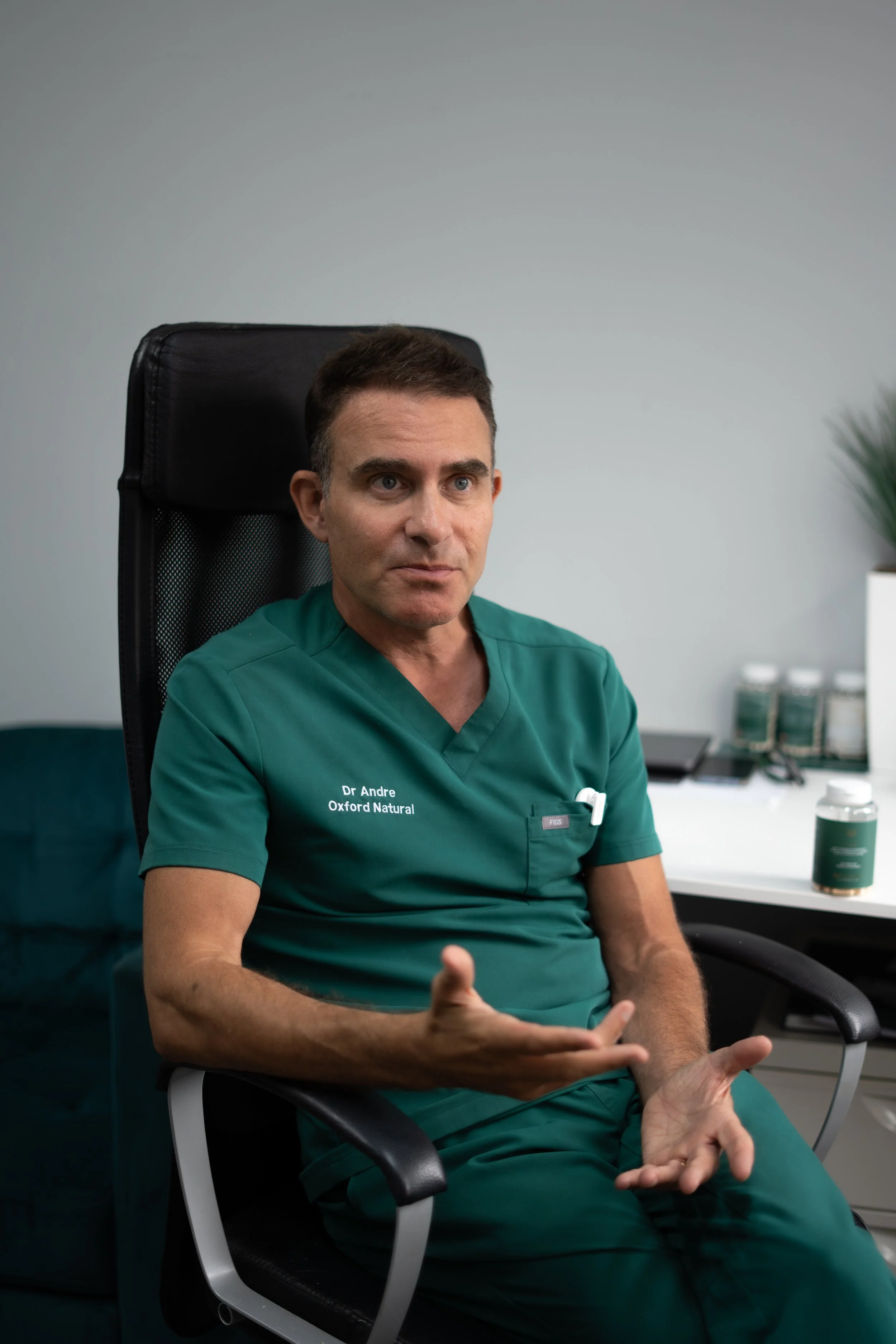 A male doctor in green scrubs sitting in an office, gesturing with his hands while speaking.