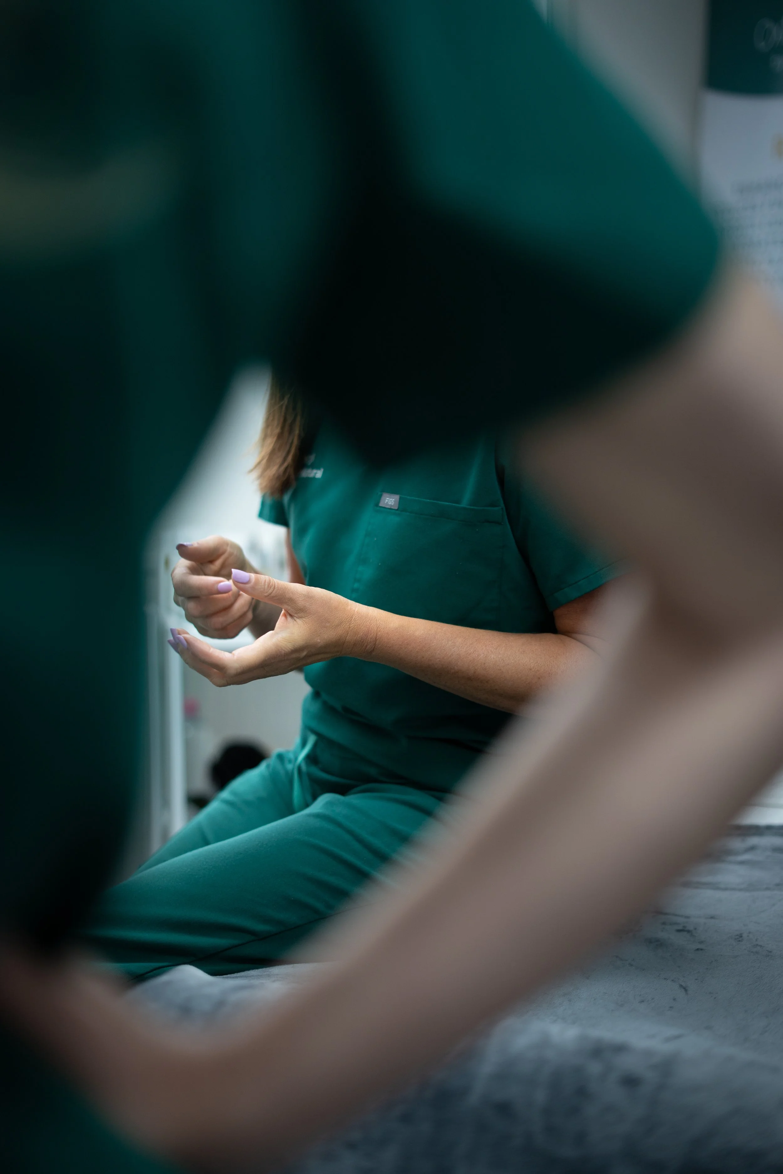 A person in green medical scrubs sitting and talking to others, with part of their body and hands visible.