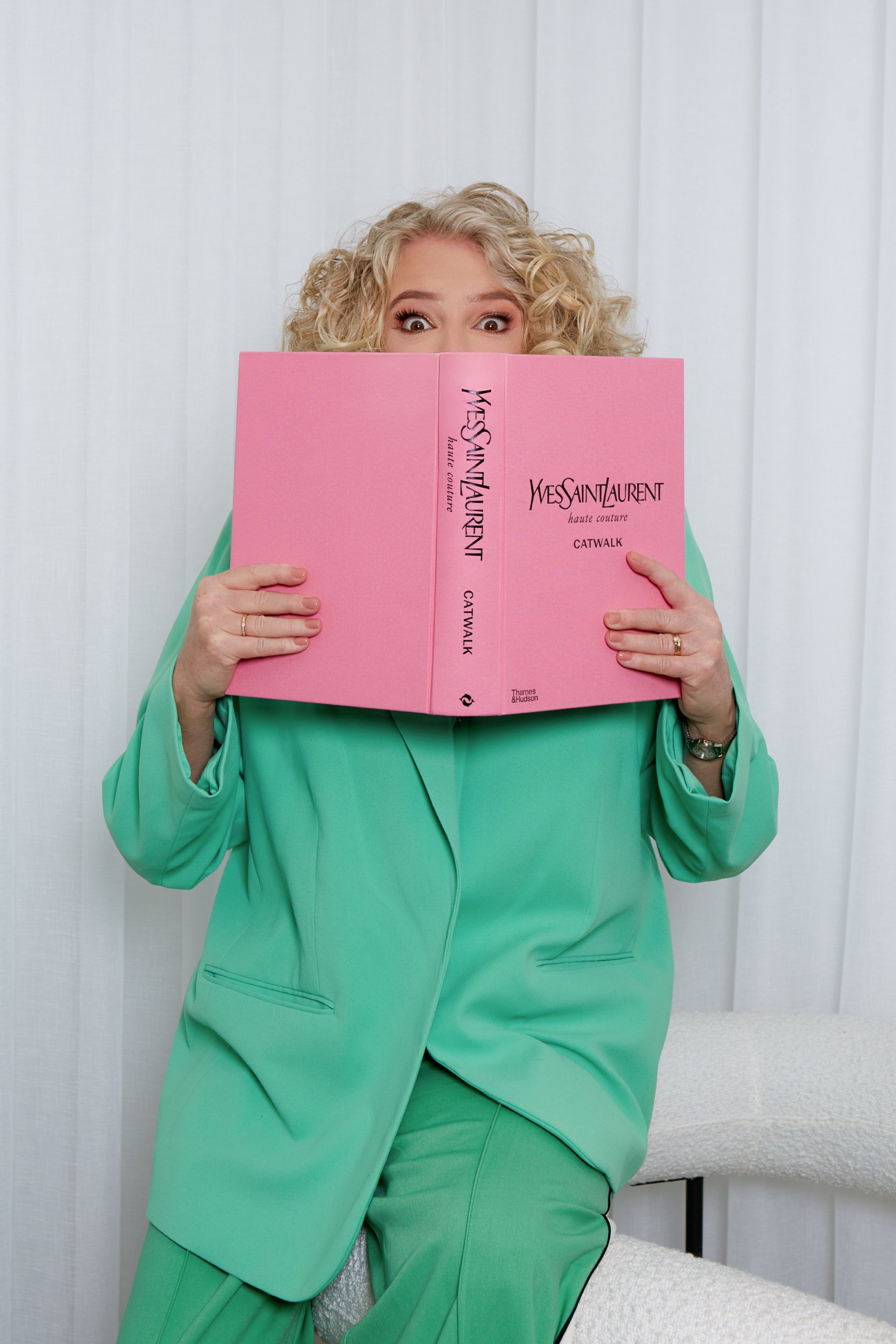 A woman with curly blonde hair wearing a green suit, holding a pink book titled 'Yves Saint Laurent Haute Couture Catwalk,' partially covering her face and looking at the camera.