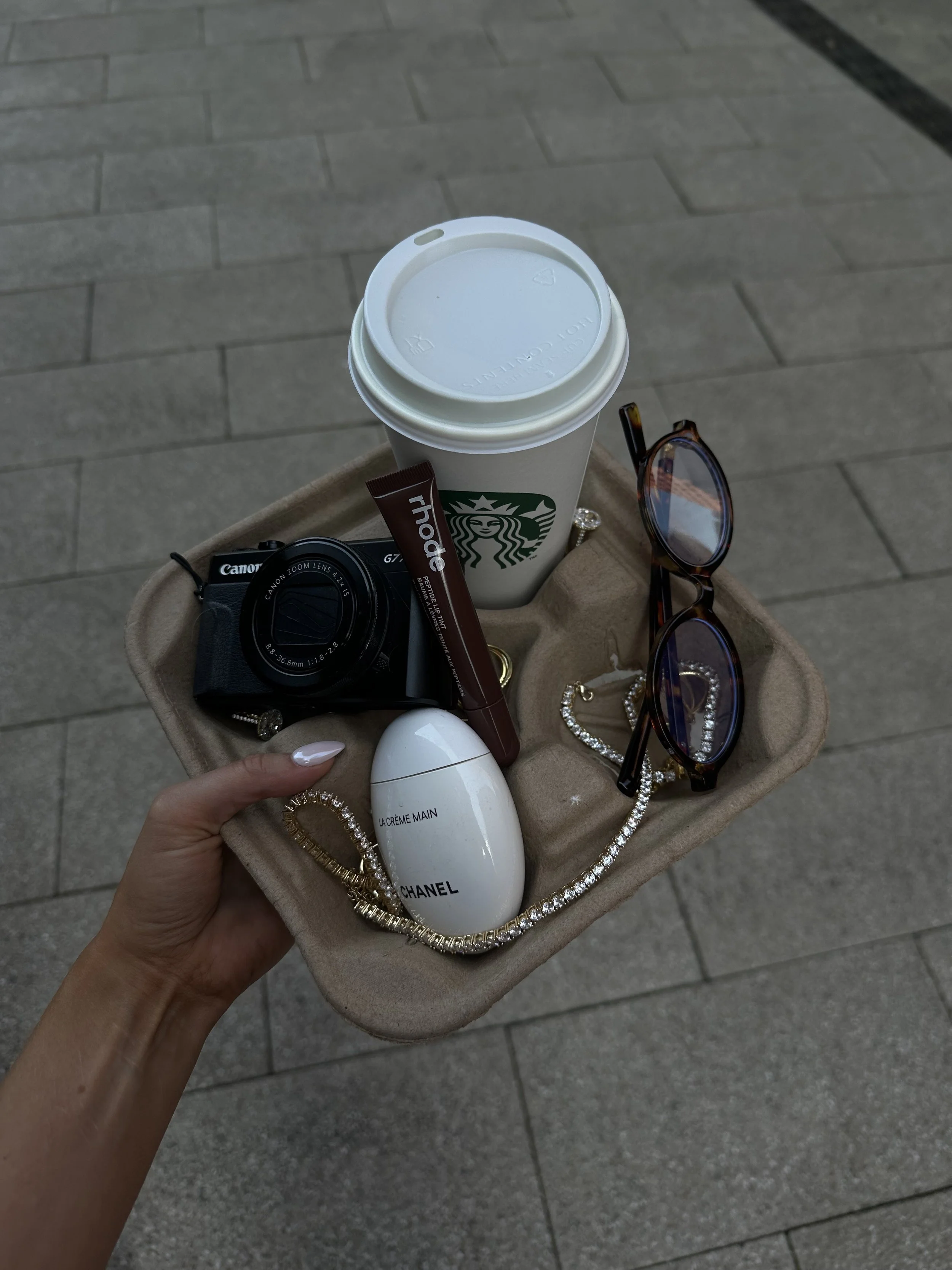 Hand holding a beige tray with a Starbucks coffee, a small camera, a Chanel hand cream, a chocolate stick, a pair of glasses, and a pearl necklace on a paved sidewalk.