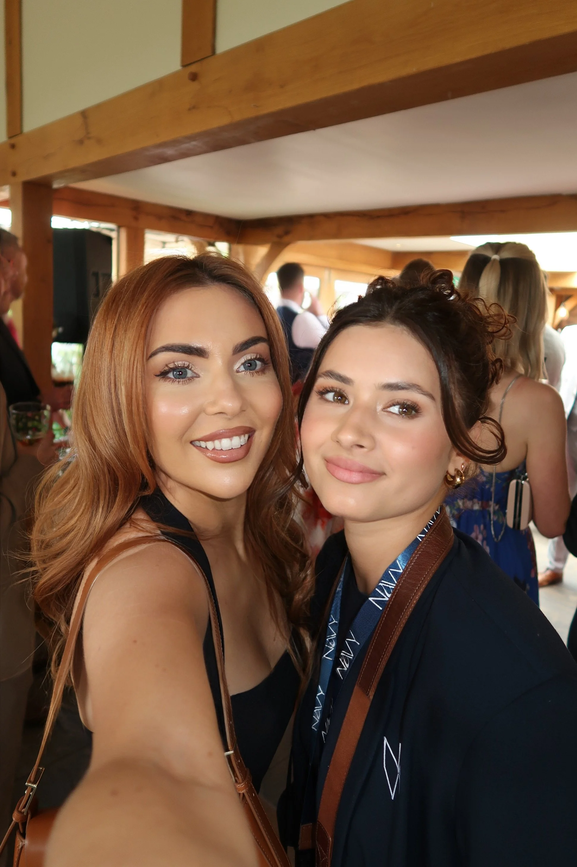 Two young women with light skin and styled hair taking a selfie at a social event in a wooden venue, smiling at the camera.