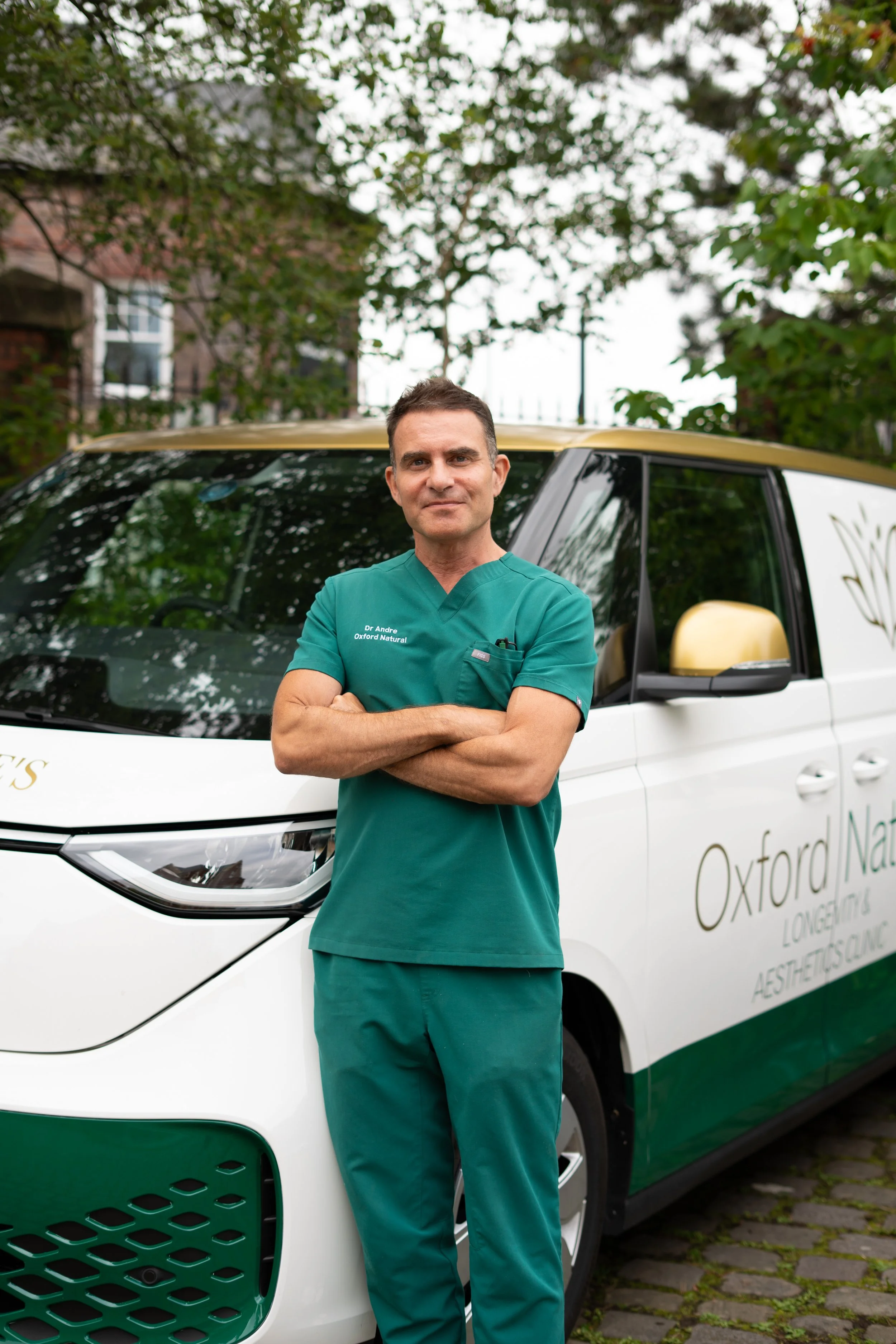 A male healthcare professional in green scrubs standing with arms crossed in front of a white vehicle that has 'Oxford Natural' written on its side, outdoors with trees and buildings in the background.