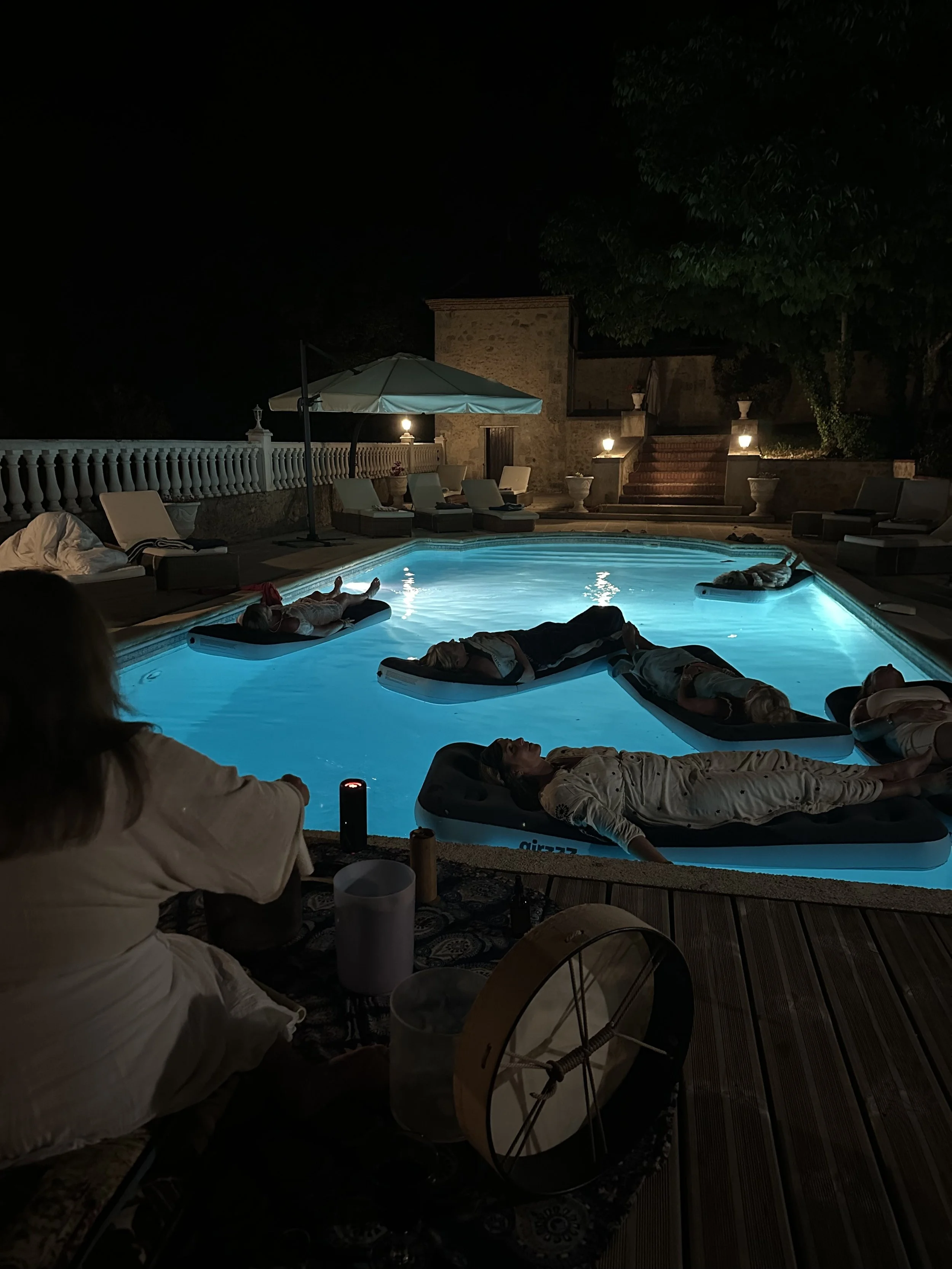 People relaxing on floating loungers in a illuminated pool at night, surrounded by outdoor furniture and decorative lighting. South bath floating on the pool in the south of france at a chateau on a wellness retreat