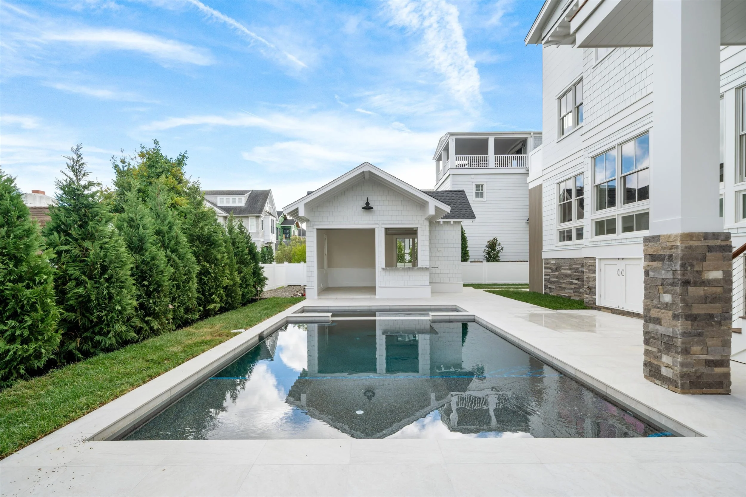 Backyard with swimming pool, white house with large windows, small white pool house, green trees, blue sky with clouds.