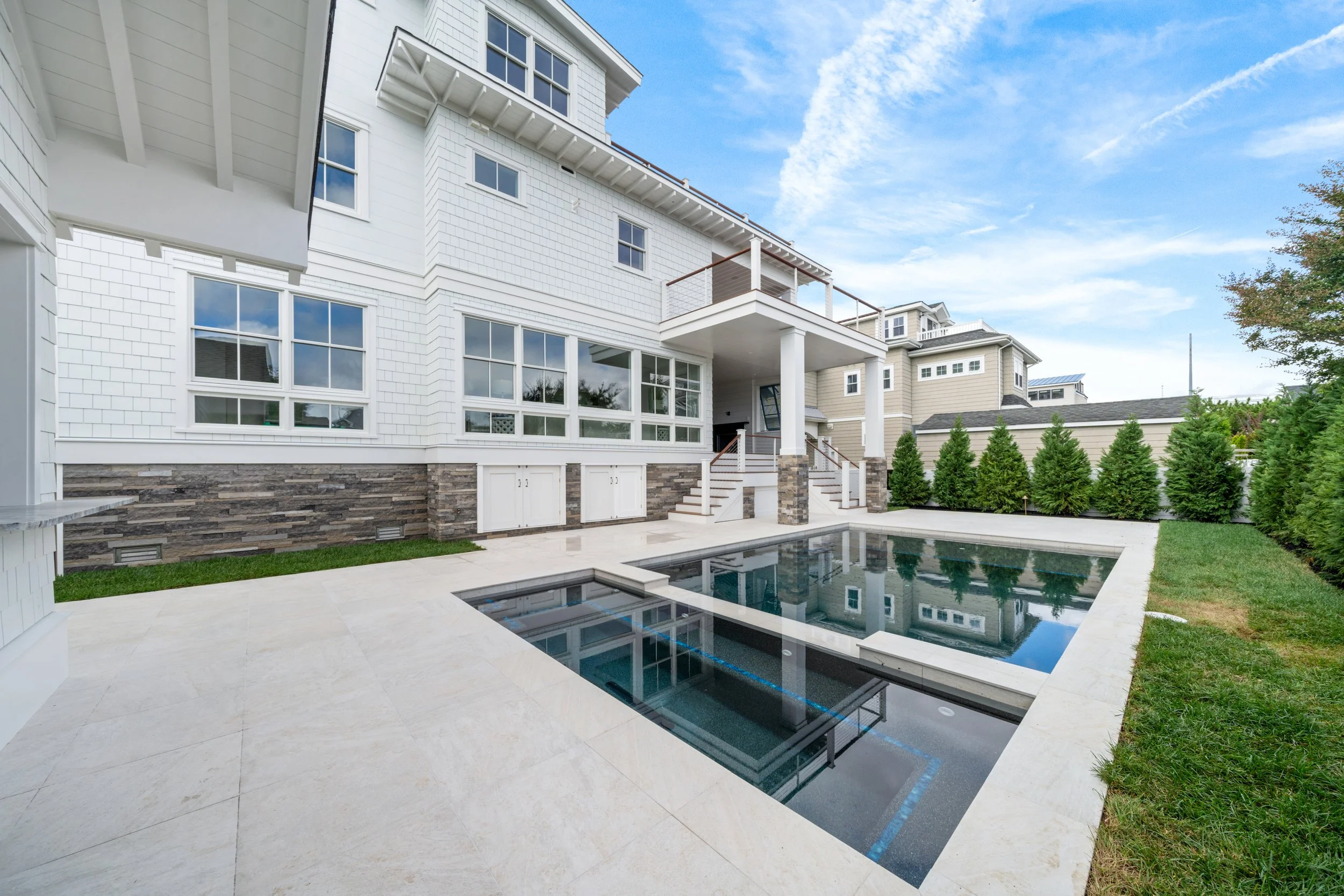 Modern backyard with a rectangular pool, white patio, and a multi-story house with large windows and a balcony, surrounded by green bushes under a blue sky.