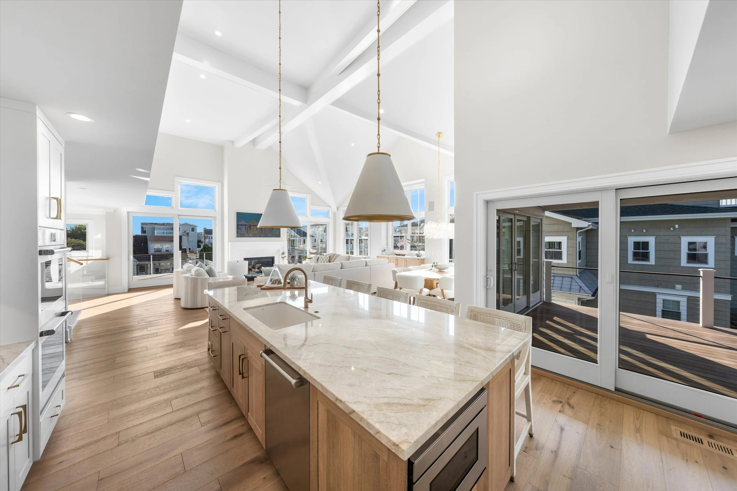 Open-concept modern kitchen with large marble island, light wood cabinets, and pendant lighting, leading to a bright living area with large windows and sliding glass doors to an outdoor deck.