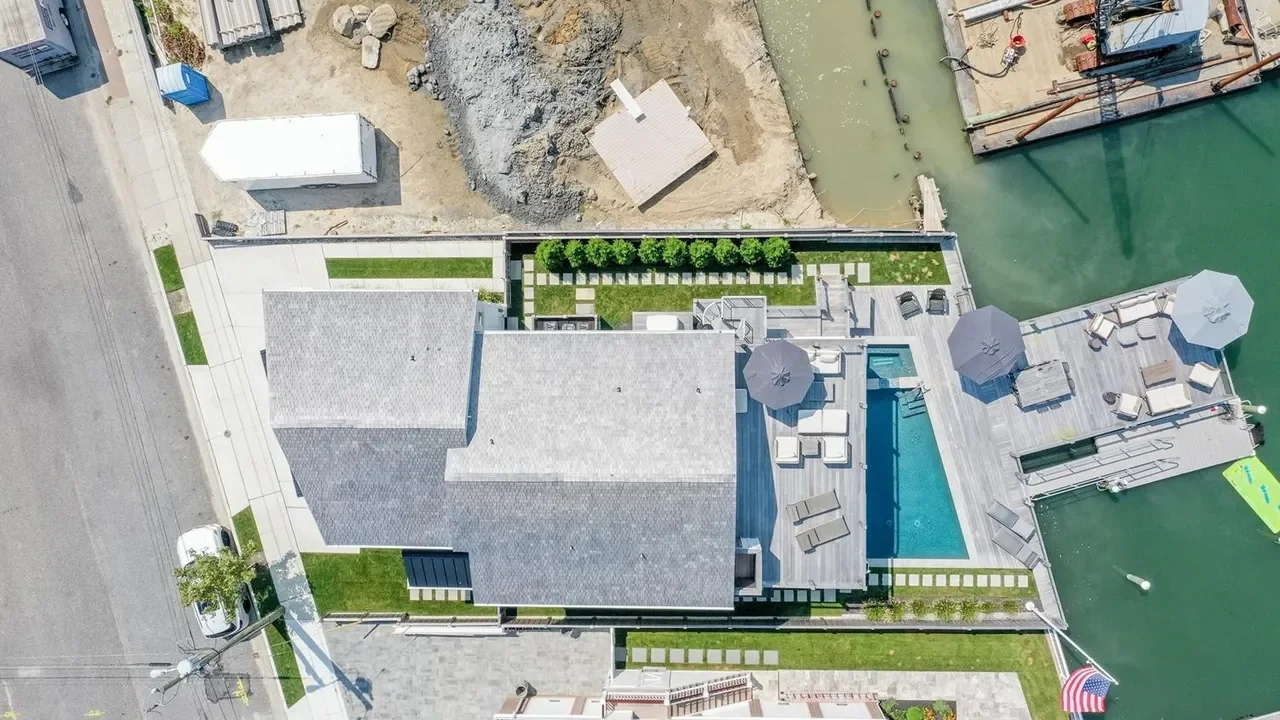 Aerial view of a modern house with a backyard pool, patio furniture, umbrellas, and a dock on a waterfront.