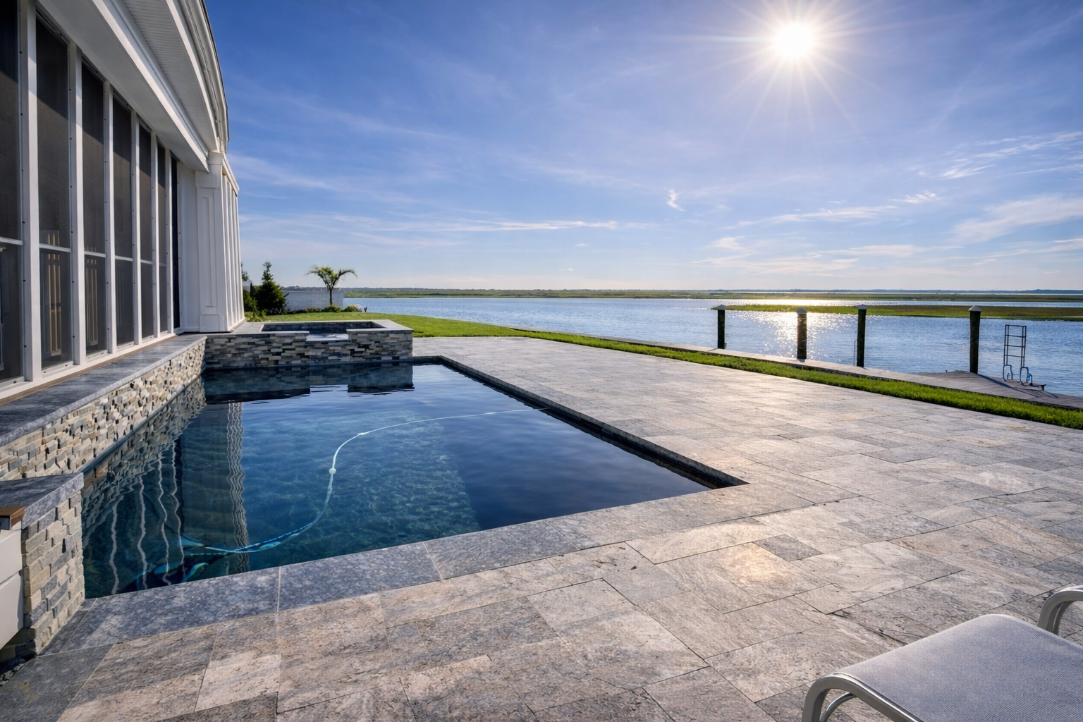 Residential backyard with a view of a lake, featuring a swimming pool, stone patio, and a chair in the foreground, under a clear sky with the sun shining.
