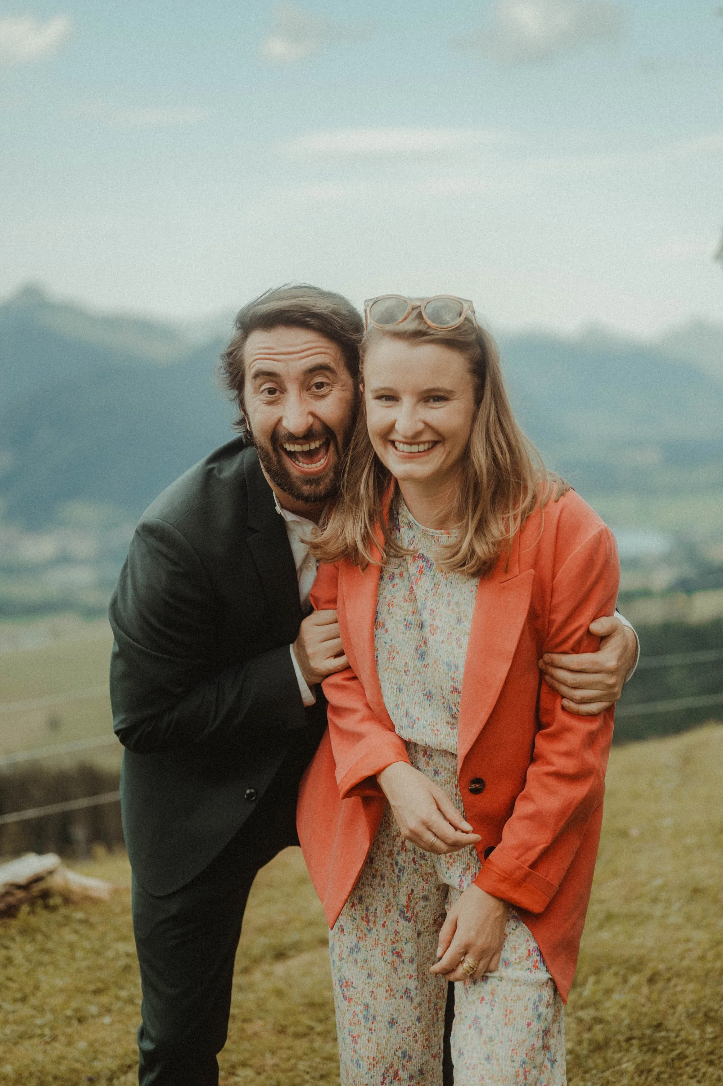 Zwei glückliche Personen im Freien vor einem Bergpanorama, eine Person im Anzug und die andere in einem roten Blazer.