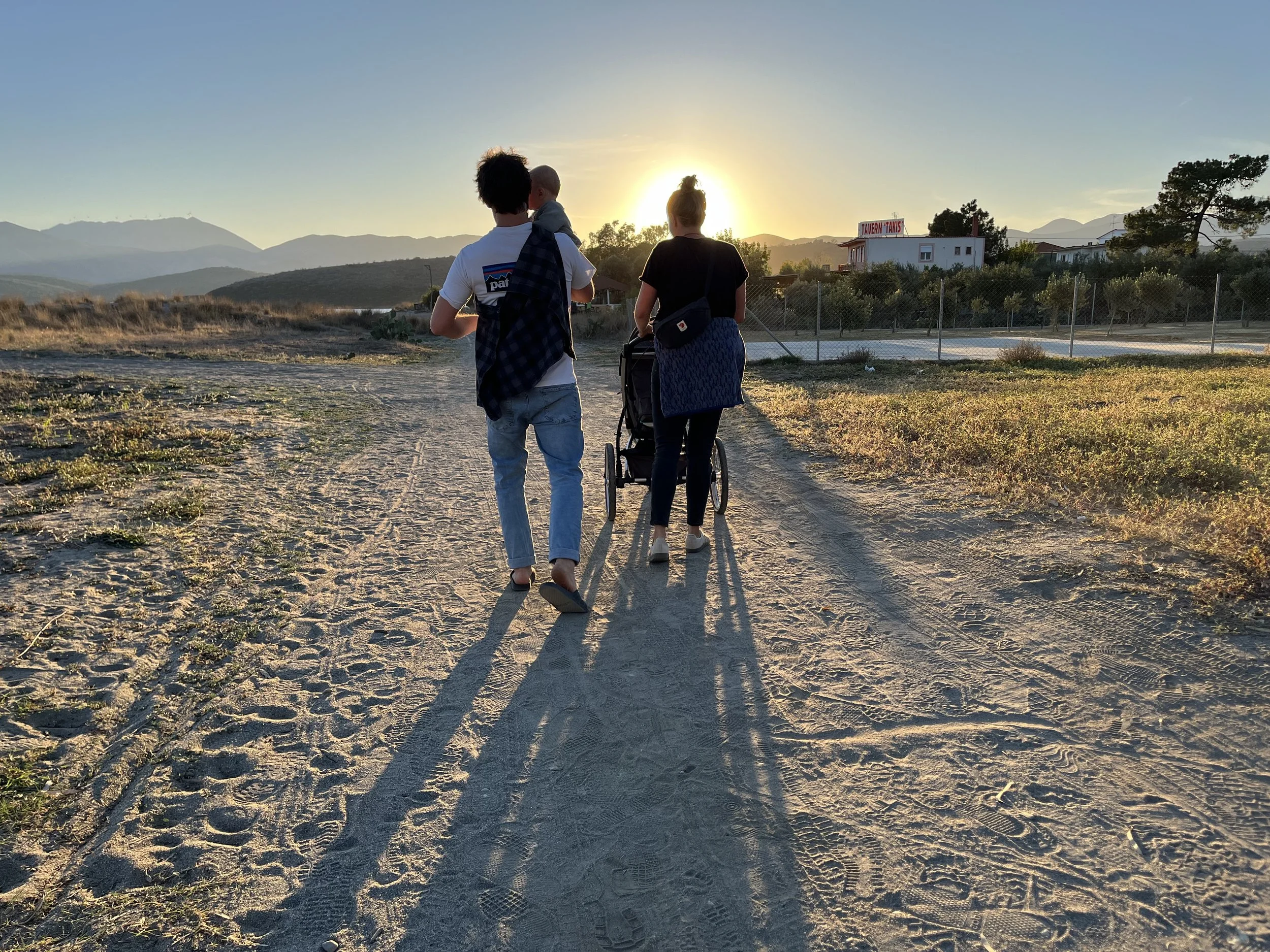 Ein Paar geht bei Sonnenuntergang auf einem Feldweg, eine Person trägt ein Kind, die andere schiebt einen Kinderwagen. Im Hintergrund sind Hügel und ein Gebäude zu sehen.