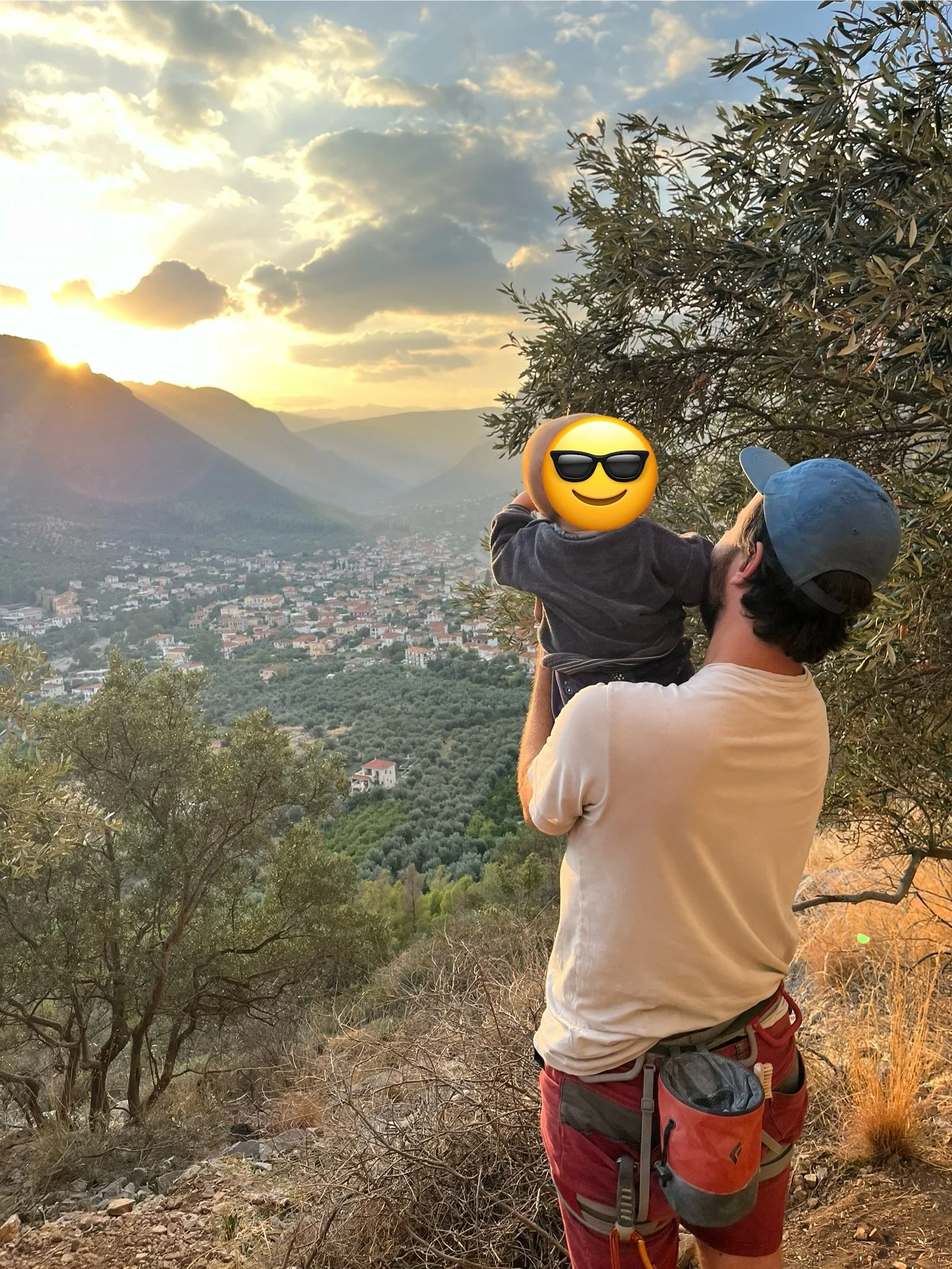 Ein Mann hebt ein Kind vor einer malerischen Berglandschaft mit einem Sonnenuntergang im Hintergrund. Beide stehen auf einem Hügel mit Bäumen und Blick auf ein Tal mit einer kleinen Stadt. Das Kind hat ein Emoji-Gesicht mit Sonnenbrille.