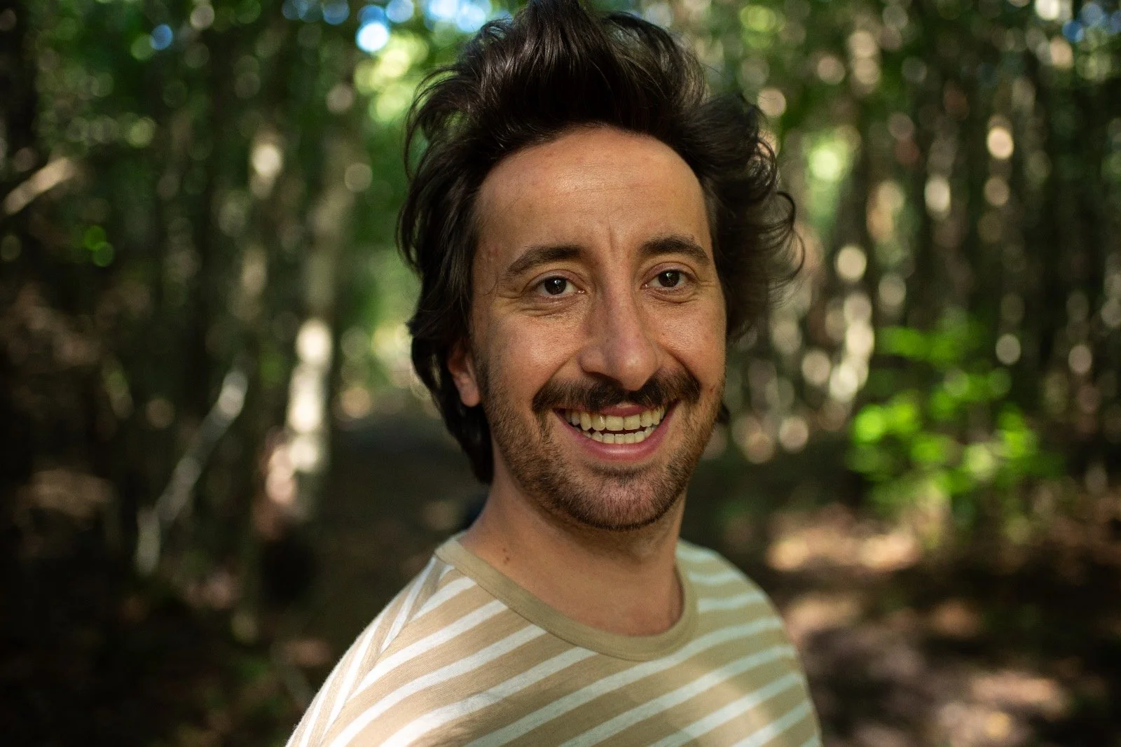 Ein Mann mit braunen Haaren und einem gestreiften T-Shirt lächelt in einem Wald.