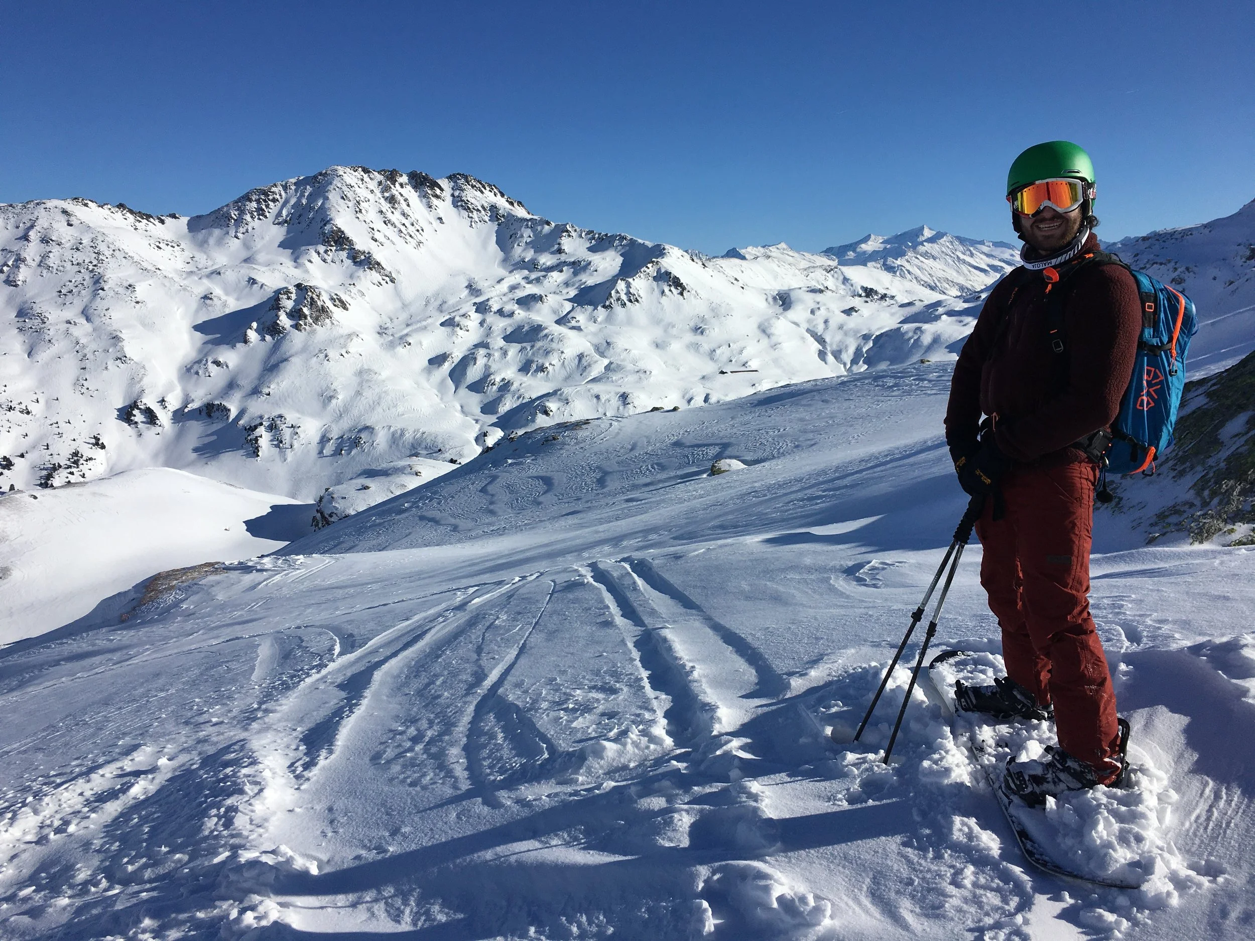 Person mit Schneeschuhen und Stöcken auf einem schneebedeckten Berg stehend, sonniger Himmel, Berglandschaft im Hintergrund.