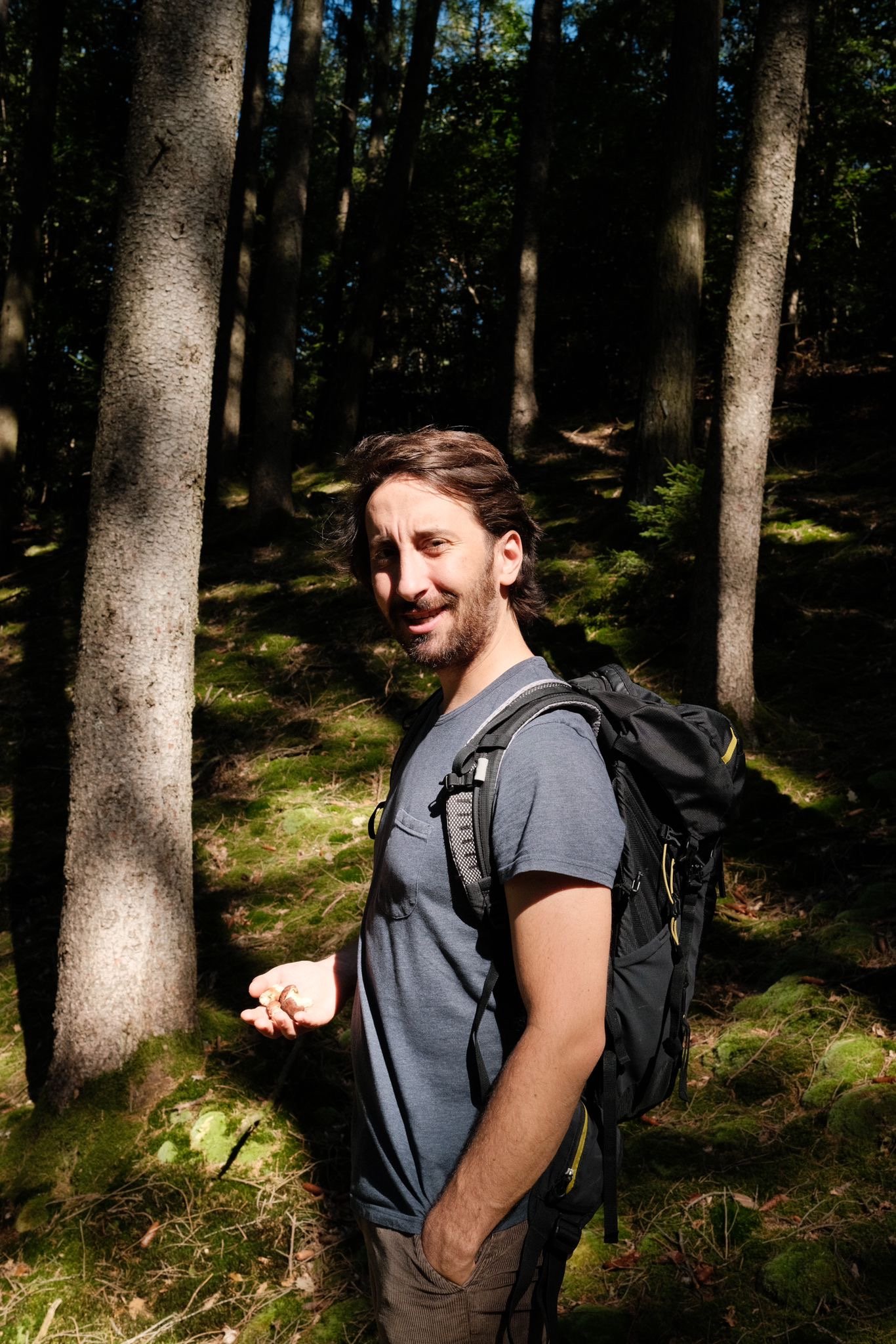 Mann mit Rucksack im Wald, Sonnenlicht durch Bäume, lächelnd.