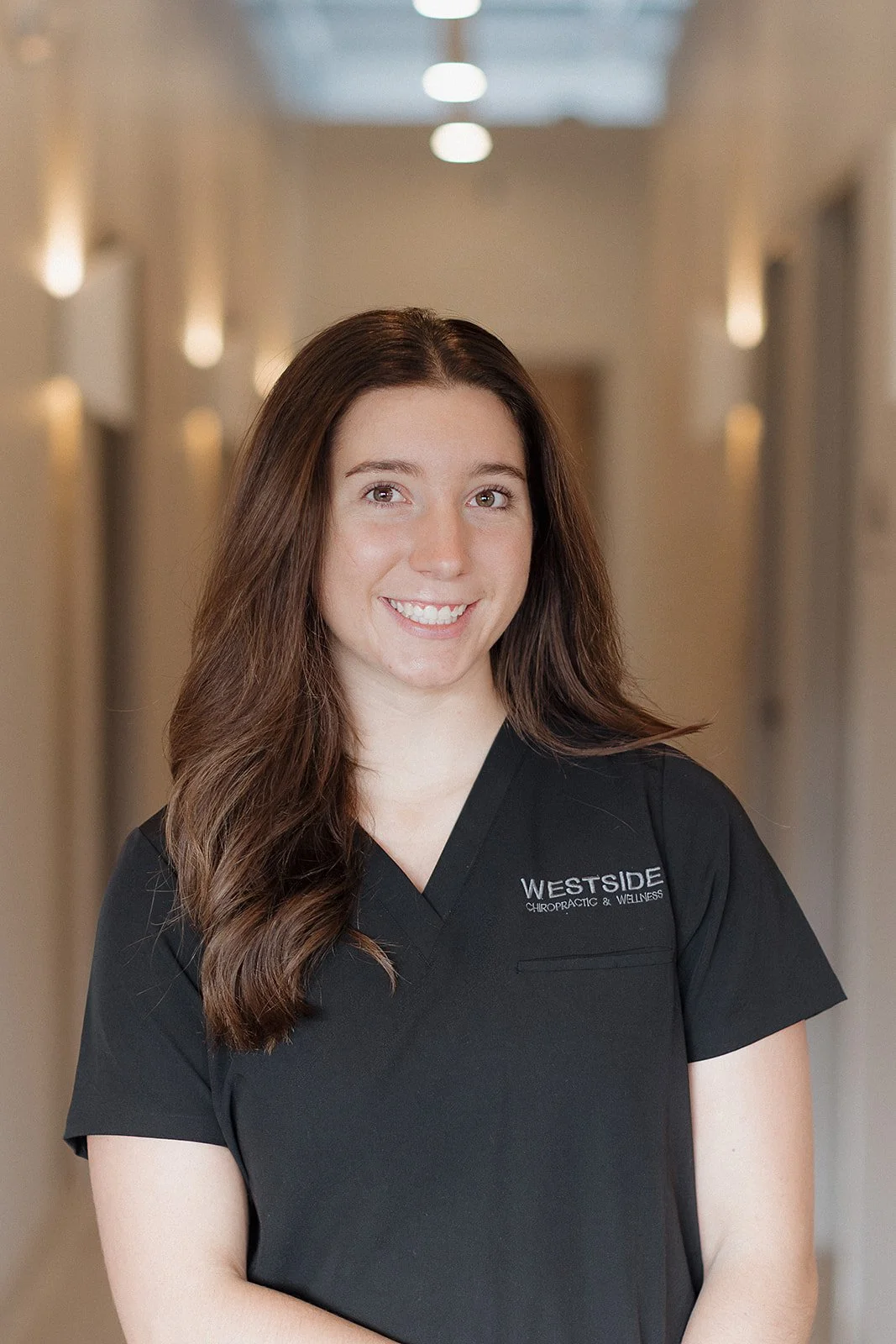 A smiling woman with long brown hair wearing a black chiropractic uniform that says "WESTSIDE CHIROPRACTIC & WELLNESS" standing in a well-lit hallway.